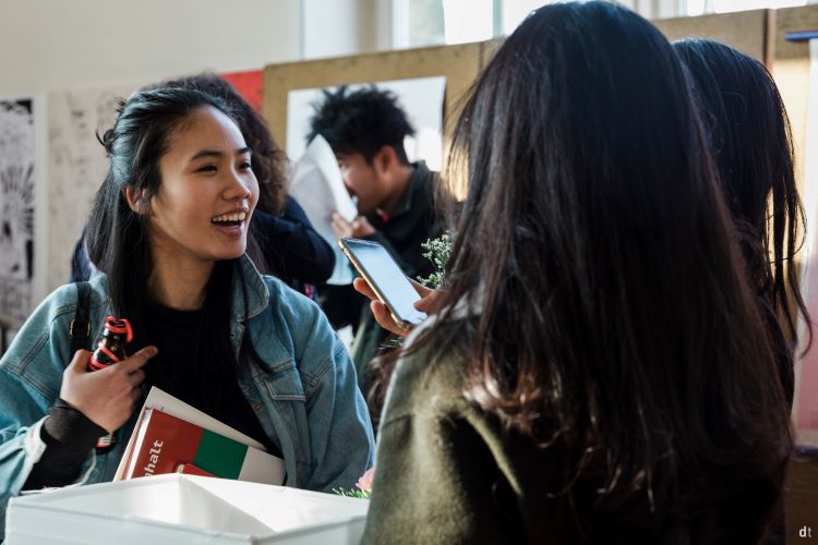 Zwei Frauen unterhalten sich und lachen, während sie ihre Smartphones halten. Eine von ihnen trägt eine Jeansjacke und hält einen Flyer. Im Hintergrund ist eine dritte Person zu sehen, die ebenfalls ein Smartphone nutzt. Die Szene wirkt locker und gesellig.