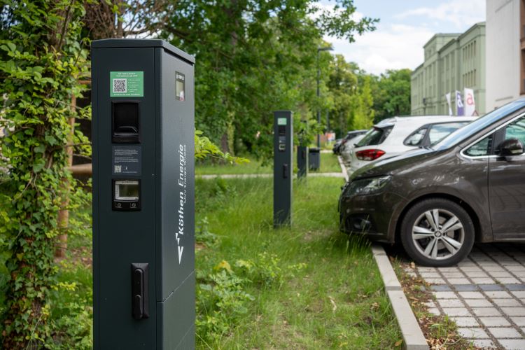 "Ein Elektroauto ist an einer Ladestation angeschlossen. Die Ladestation hat ein Display und verschiedene Anschlüsse. Im Hintergrund sind weitere Ladestationen und geparkte Autos zu sehen. Die Szene spielt auf einem bewachsenen Parkplatz."