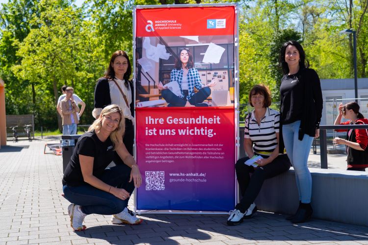 "Vier Personen stehen vor einem Plakat der Hochschule Aalen. Das Plakat betont die Bedeutung der Gesundheit mit dem Text 'Ihre Gesundheit ist uns wichtig' und zeigt eine Person im Sitzen. Die Website www.hs-aalent.de/gesundheit-hochschule wird erwähnt."