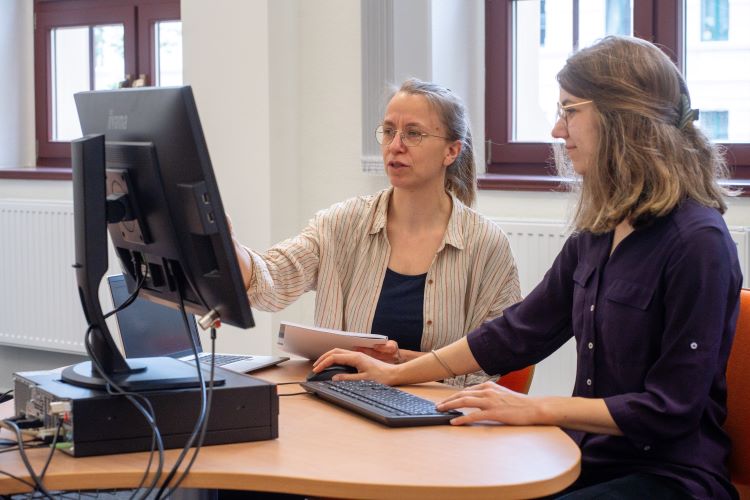 Two women are working together at a computer station. One woman is seated, using the keyboard, while the other stands beside her, pointing at the screen and providing guidance. They appear to be engaged in a collaborative task or discussion.