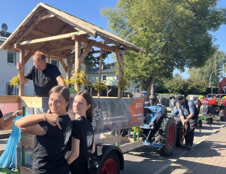 "Zwei Frauen stehen lächelnd vor einem Traktor mit Anhänger. Auf dem Anhänger befindet sich eine Holzhütte mit einem Mann, der Blumen hält. Im Hintergrund sind weitere Traktoren und Menschen zu sehen. Die Szene wirkt wie ein Fest oder eine Parade."