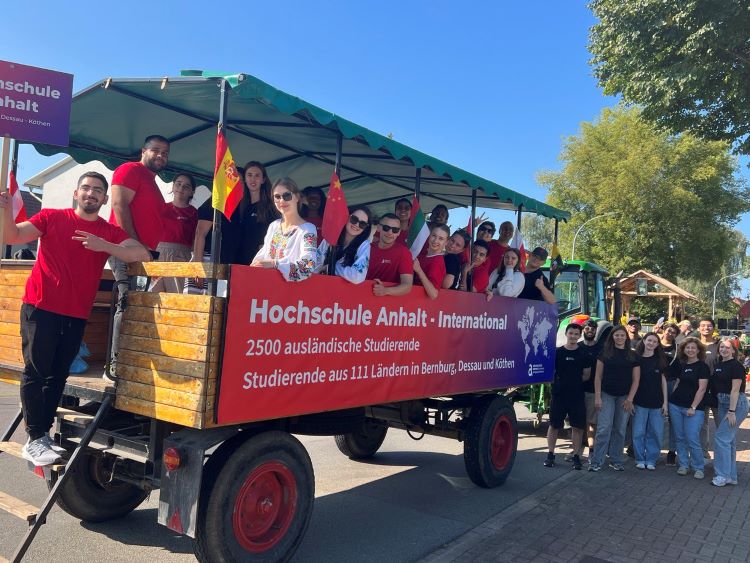 Ein Gruppenbild von Studierenden der Hochschule Anhalt - International. Sie stehen auf und neben einem Anhänger, der die Information trägt, dass 2500 ausländische Studierende aus 111 Ländern in Bernburg, Dessau und Köthen studieren. Die Studierenden halten Flaggen und wirken fröhlich.