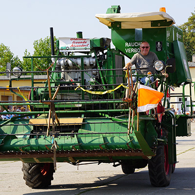 A man is operating a large green agricultural machine, likely a forage harvester, during a parade or public event. The machine has a sign on it, and the man is wearing a hat and sunglasses. The background shows a few spectators and buildings.