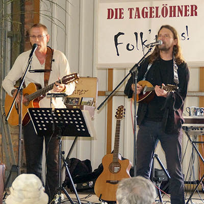 Two men are performing on stage with acoustic guitars. One is singing into a microphone while the other plays guitar. They are part of a folk music group named "Die Tagelöhner," as indicated by the banner behind them. The audience is watching attentively.
