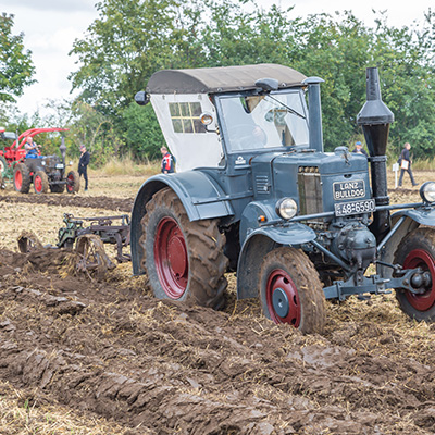 An old blue tractor with red wheels is plowing a muddy field. The tractor has a white canopy and is being operated by a person. In the background, there are other tractors and people observing the activity in a rural setting with trees.