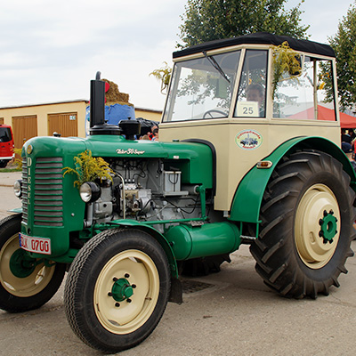 A vintage green and beige tractor with large rear tires and smaller front tires is parked on a paved area. The tractor is adorned with yellow flowers on its front and side. The license plate reads "AK 6700". The background shows some buildings and trees.