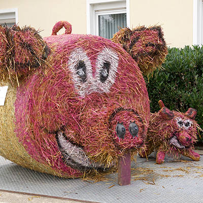 Two large, colorful sculptures of pigs made from straw and other materials are displayed outdoors. The larger pig has a big smile, while the smaller pig has a more neutral expression. Both sculptures have exaggerated features and are placed on a paved surface.