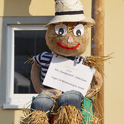 A scarecrow figure wearing a hat, striped shirt, and jeans, holding a sign that reads "Kita 'Zwergenland' Calbe/Saale" and mentions "Trigger: LH Bördeland gGmbH." The scarecrow is made of straw and has a friendly face with a red smile and large white eyes.