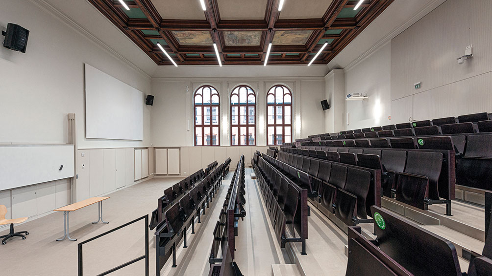 Ein leerer Hörsaal mit mehreren Reihen schwarzer Stühle, die zu einem Podium mit einem Whiteboard ausgerichtet sind. Große Bogenfenster lassen Licht herein, und die Decke hat eine Holzvertäfelung mit eingebauten Lichtern. Der Raum wirkt sauber und modern.