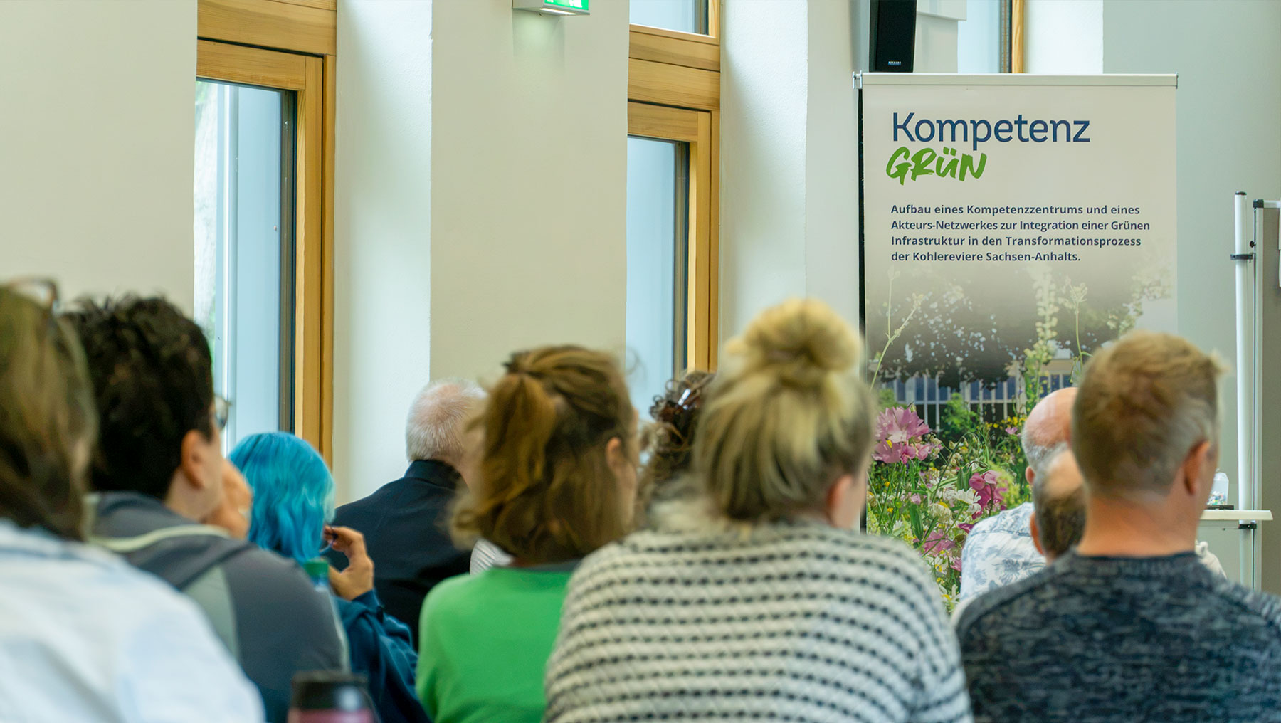Gruppe von Menschen in einem Raum, die sich einem Plakat mit dem Titel Kompetenz Grün zuwendet. Das Plakat zeigt eine grüne Landschaft mit Pflanzen und bezieht sich auf Kompetenzzentren und Transformation im Bereich Grünflächen und Nachhaltigkeit.