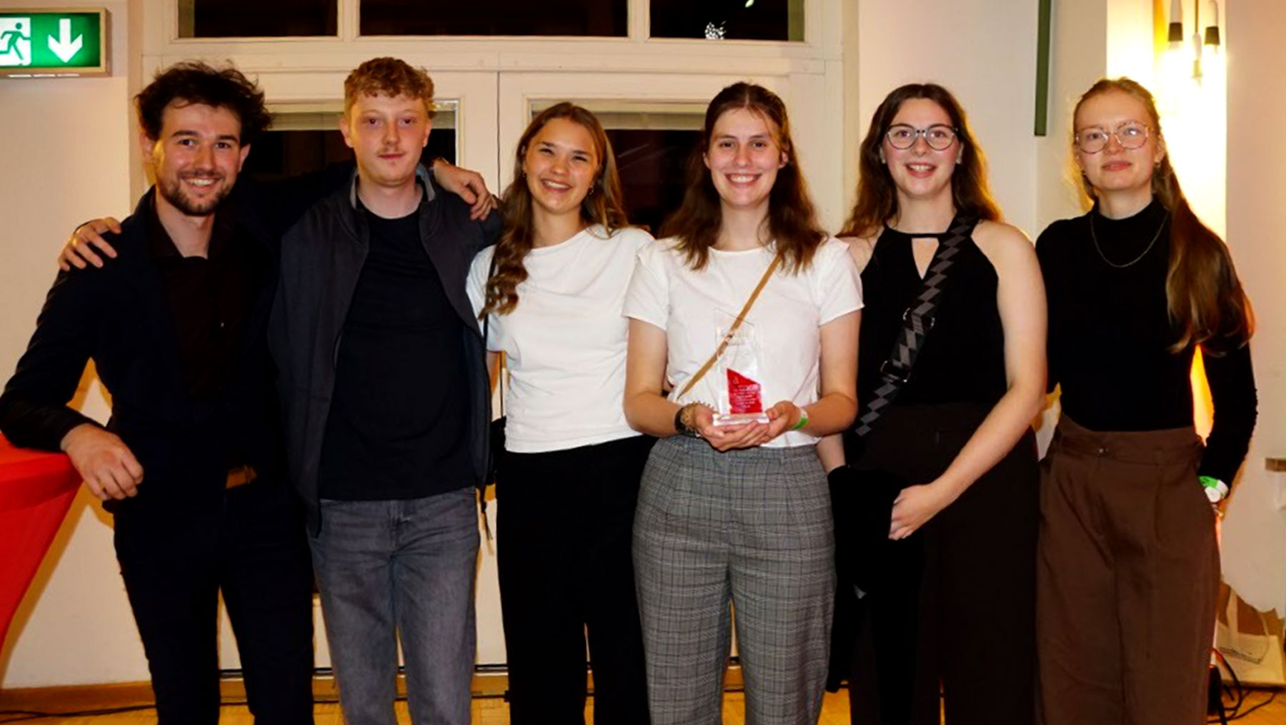 Fünf Personen stehen lächelnd beieinander, eine hält einen kleinen Pokal in Form eines Herzens mit einem Band. Die Gruppe wirkt zufrieden und feiert gemeinsam einen Erfolg.