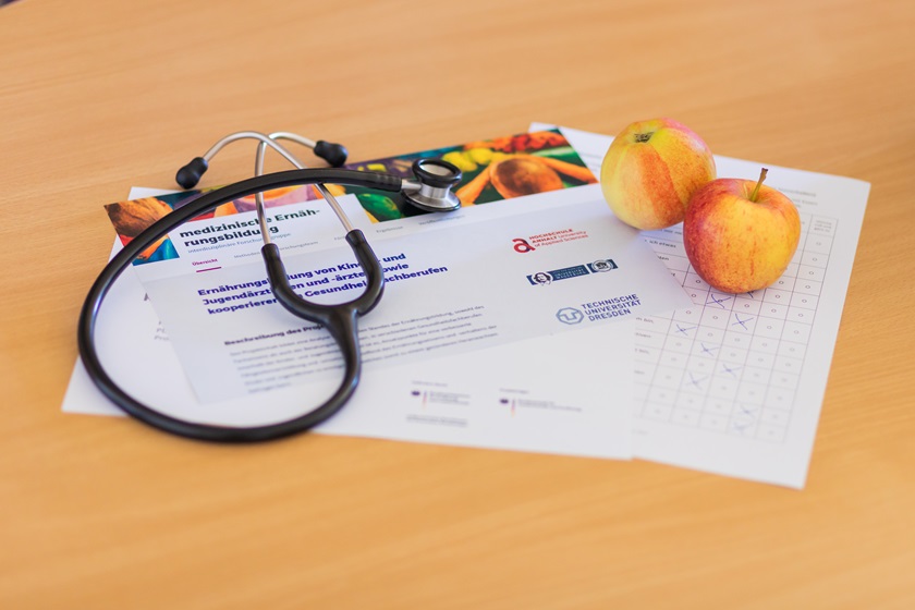 Stethoskop liegt auf einem Informationsblatt zur medizinischen Ernährung mit Hinweisen zu gesunder Kost, daneben zwei Äpfel als Symbol für gesunde Lebensmittel.