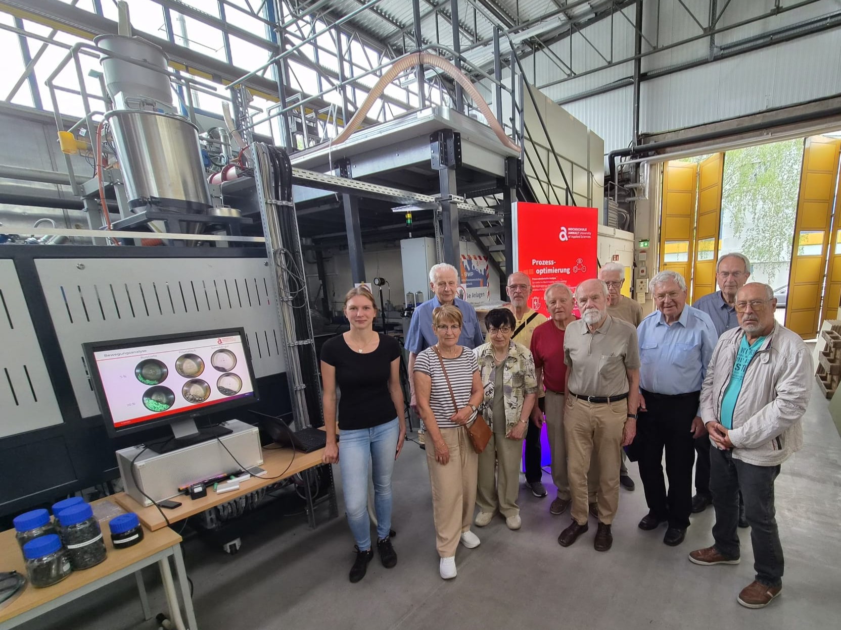 Gruppe von Menschen, darunter eine jüngere Frau und mehrere ältere Herren und Damen, stehen vor einer großen industriellen Maschine in einer Fabrikhalle und lächeln in die Kamera. Die Maschine hat ein Display mit medizinischen Bildschirminhalten.