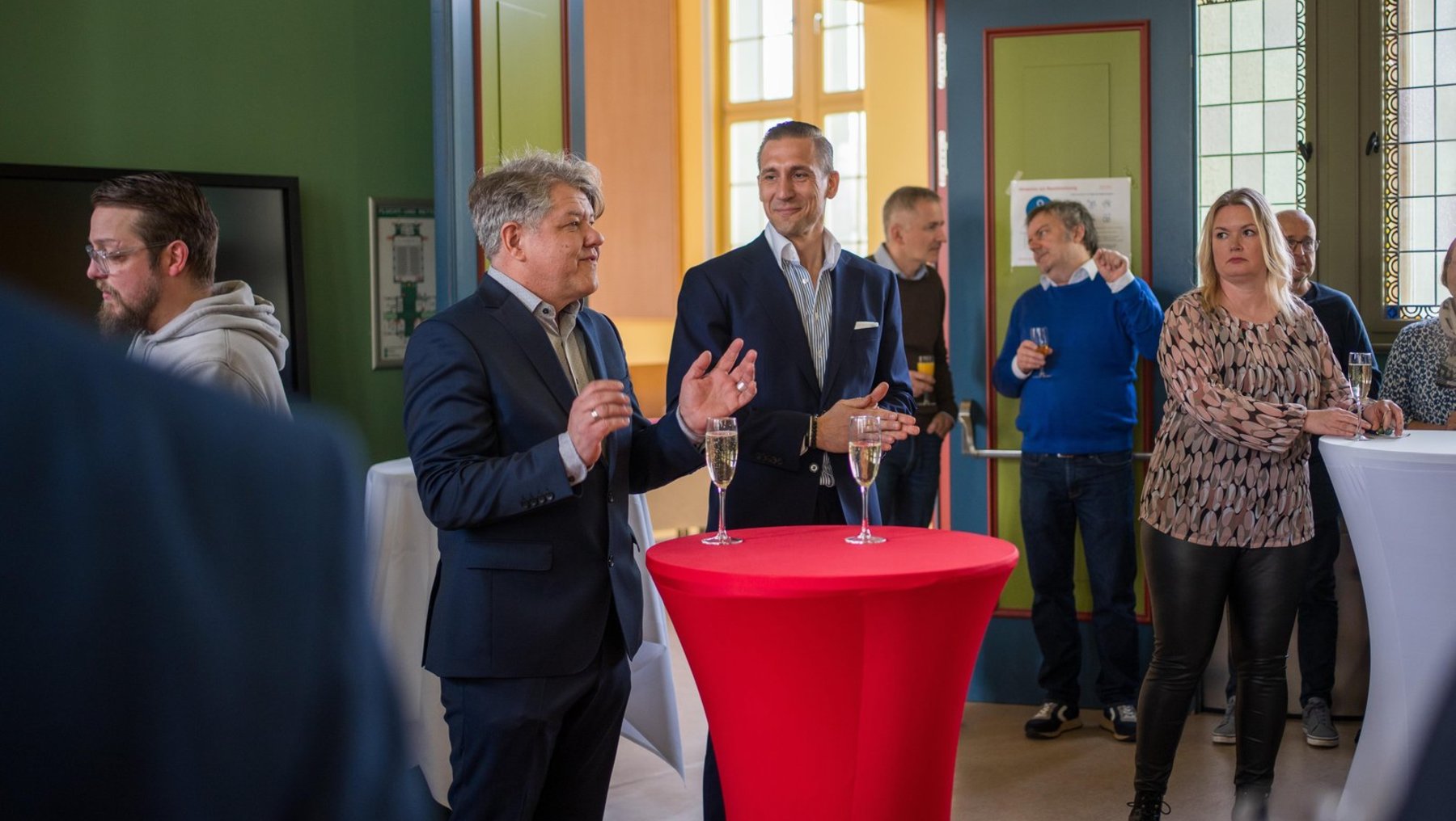 Gruppe von Menschen bei einer kleinen Feier oder Veranstaltung, die Sektgläser in der Hand halten. Zwei Herren im Vordergrund klatschen, während andere Personen im Hintergrund stehen und sich unterhalten. Ein roter runder Tisch mit Sektflasche und Gläsern steht zentral.