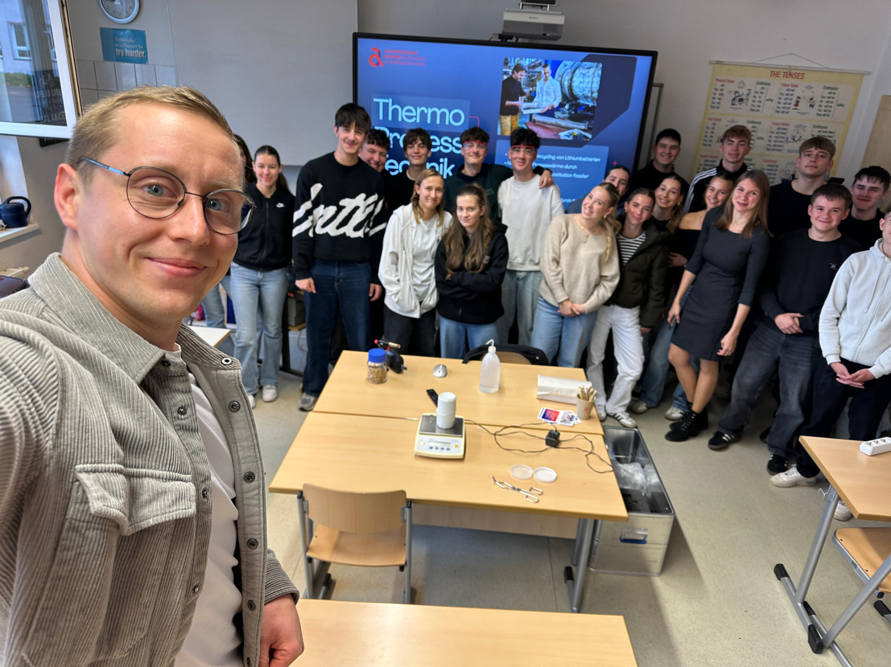 Gruppe von etwa 16 Personen, darunter ein Mann vorne links, steht lächelnd in einem Klassenzimmer mit Tischen und Experimentiermaterialien wie Thermometer und Messgeräte. Im Hintergrund ein Bildschirm mit dem Text „Thermo-Elemente“.