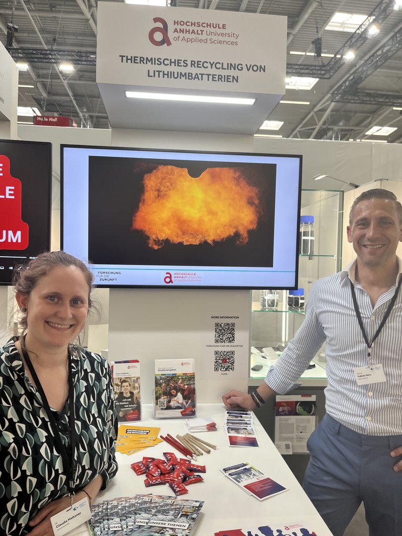 Zwei Personen stehen lächelnd an einem Infostand der Hochschule Anhalt zum Thema „Thermisches Recycling von Lithiumbatterien“. Im Hintergrund ist eine große Flamme auf einem Bildschirm zu sehen, auf dem Tisch liegen Informationsmaterialien und Batterieproben.