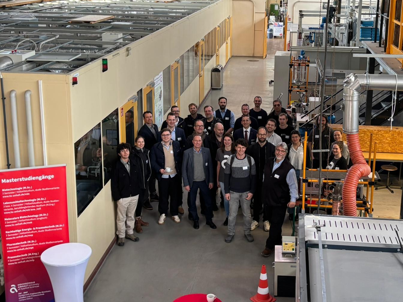 Gruppe von etwa 20 Personen steht lächelnd in einer Industriehalle mit Produktionsanlagen, darunter Rohrleitungen und Maschinen. Im Hintergrund ein Schild mit „Masterstudiengang“.