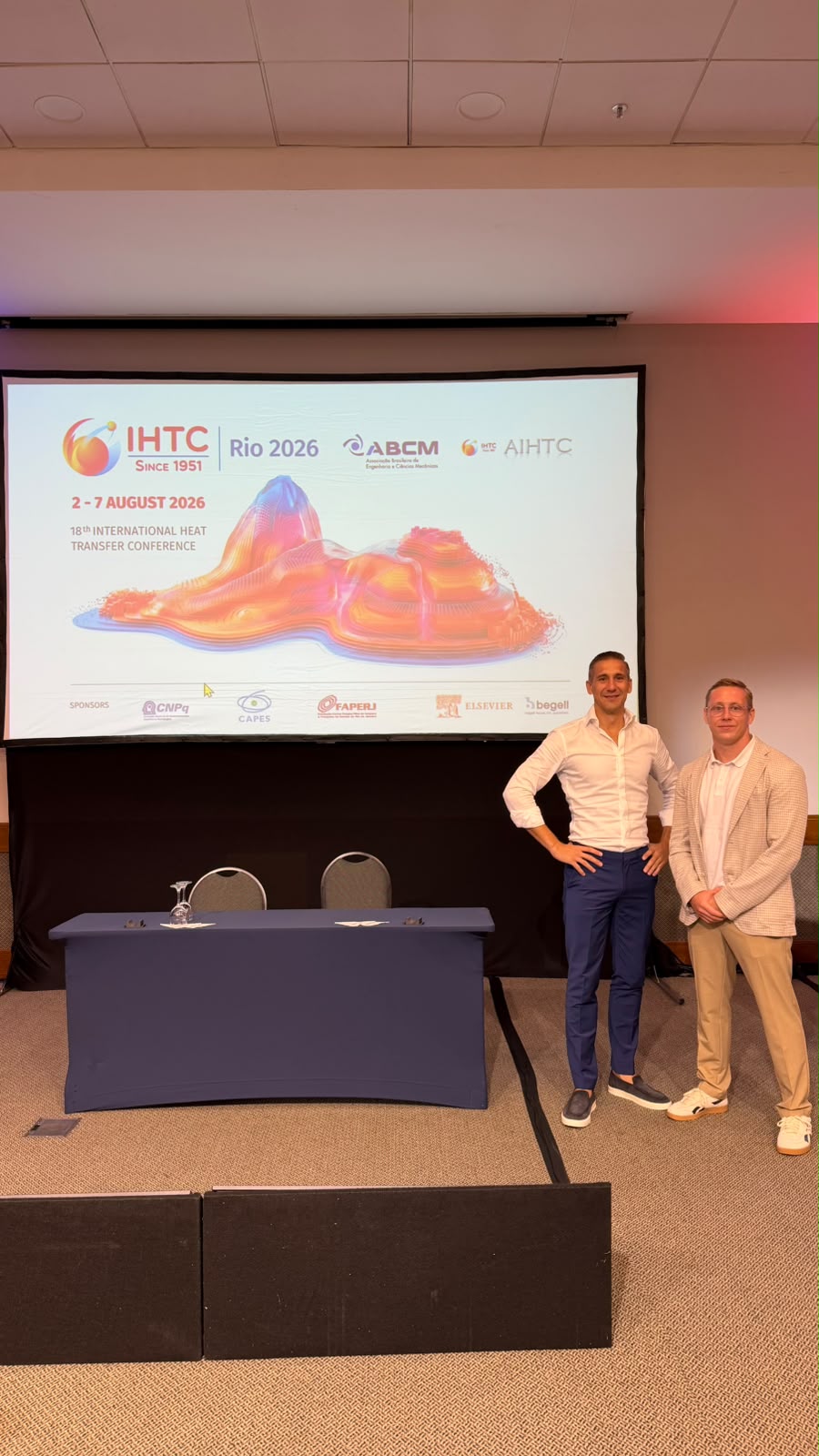 Zwei Männer stehen auf einer Bühne mit einem großen Bildschirm im Hintergrund, der die IHTC-Konferenz 2026 in Rio de Janeiro vom 2. bis 7. August 2026 ankündigt. Auf dem Bildschirm sind bunte Sneaker und Logos verschiedener Partner zu sehen.