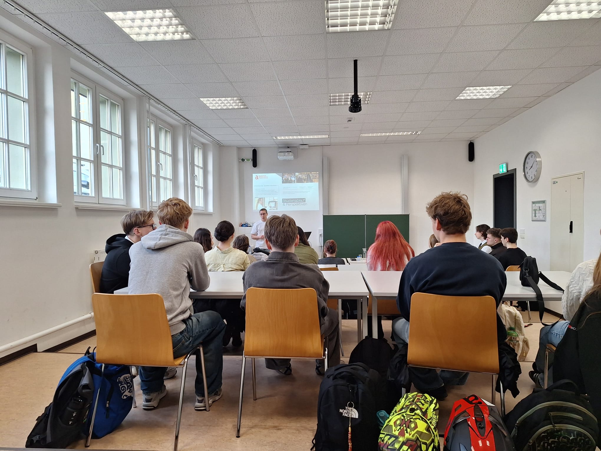 Ein Klassenzimmer mit mehreren Schülern, die an Tischen mit Stühlen sitzen und einer Person, die vorne an einer Tafel steht und eine Präsentation hält. Im Hintergrund ist ein Beamerbild an der Wand zu sehen.