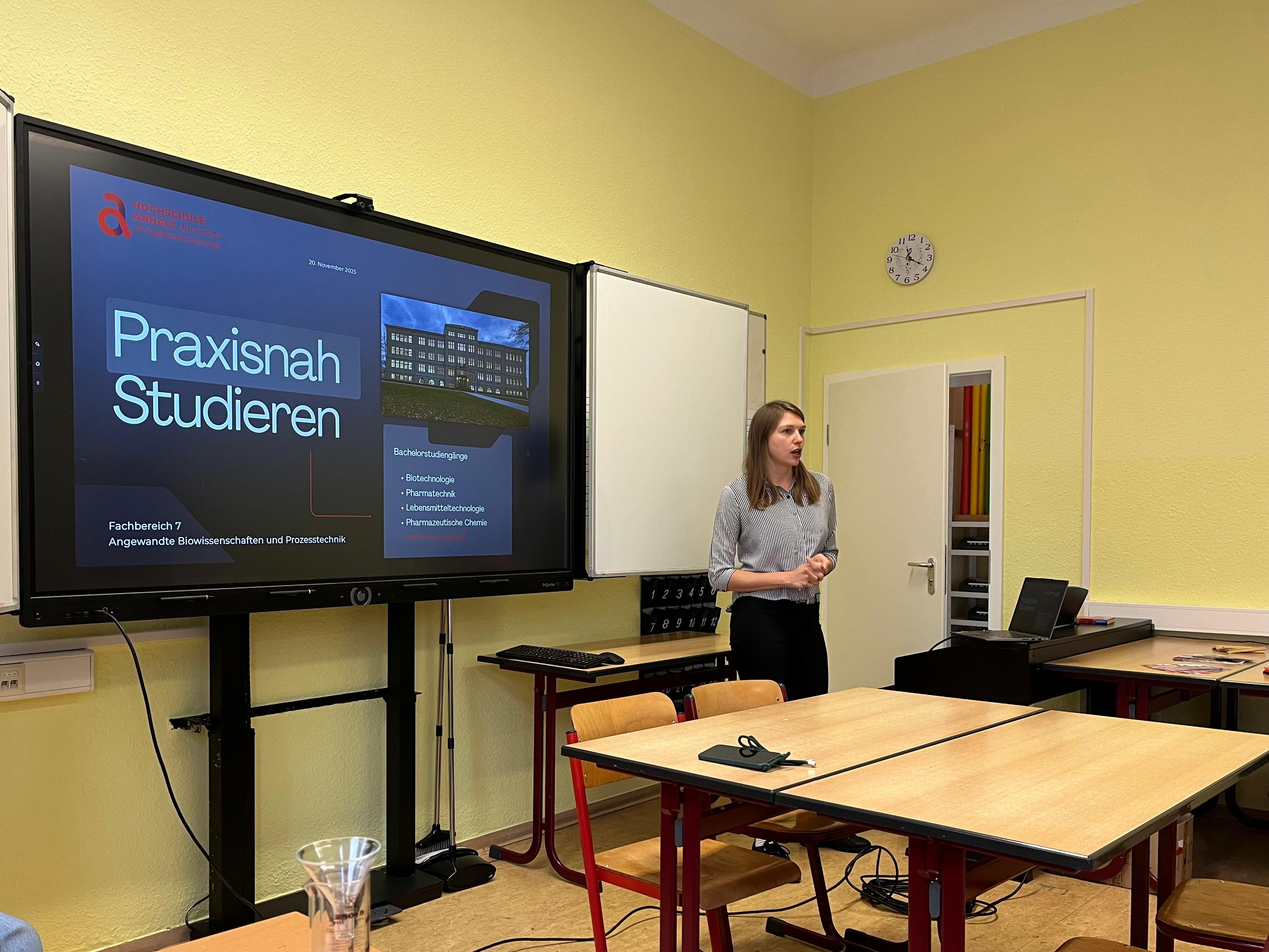 Eine Person steht in einem Seminarraum und präsentiert vor einem großen Bildschirm mit der Aufschrift „Praxisnah Studieren“ sowie Unterpunkten zu Studieninhalten wie „Angewandte Informatik und Praxissemester“. Im Hintergrund sind Tische mit Stühlen und eine Tür sichtbar.