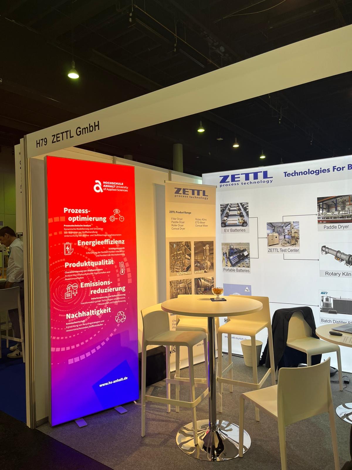 Messe-Stand der Firma ZETTL GmbH mit Fokus auf Nachhaltigkeit und Effizienz. Links ein Banner mit den Schwerpunkten Prozessoptimierung, Energieeffizienz, Produktivität, Emissionsreduzierung und Nachhaltigkeit. Im Hintergrund Infotafeln zu Technologien und Anwendungen wie Public Display und Roboter-Kinematik.