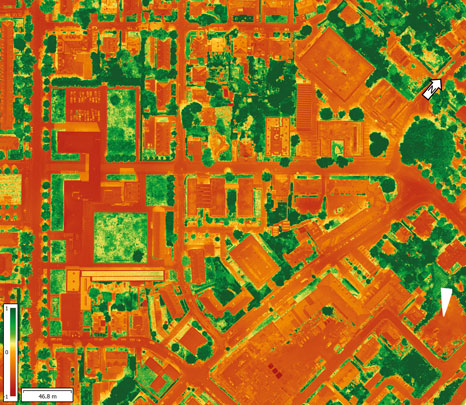 Luftbild einer Stadt mit farbkodierter Infrarotdarstellung, das Gebäude in warmen Tönen (rot/orange) und Grünflächen sowie Vegetation in grünen und dunklen Tönen zeigt. Zwei Pfeile markieren spezifische Bereiche im Bild.