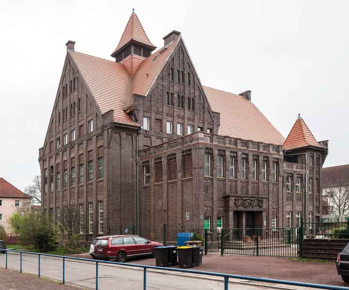 Großes historisches Backsteingebäude mit steilem Satteldach, Zwerchhäusern und symmetrischem Fensteraufbau, vermutlich ein ehemaliges Schul- oder Verwaltungsgebäude. Vor dem Eingang steht ein roter Kleinwagen.