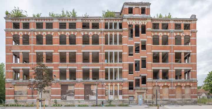 Verfallenes, mehrstöckiges Backsteingebäude mit vielen leeren, zerstörten Fensteröffnungen und fehlenden Türen, umgeben von einer leeren Straße und spärlicher Vegetation.