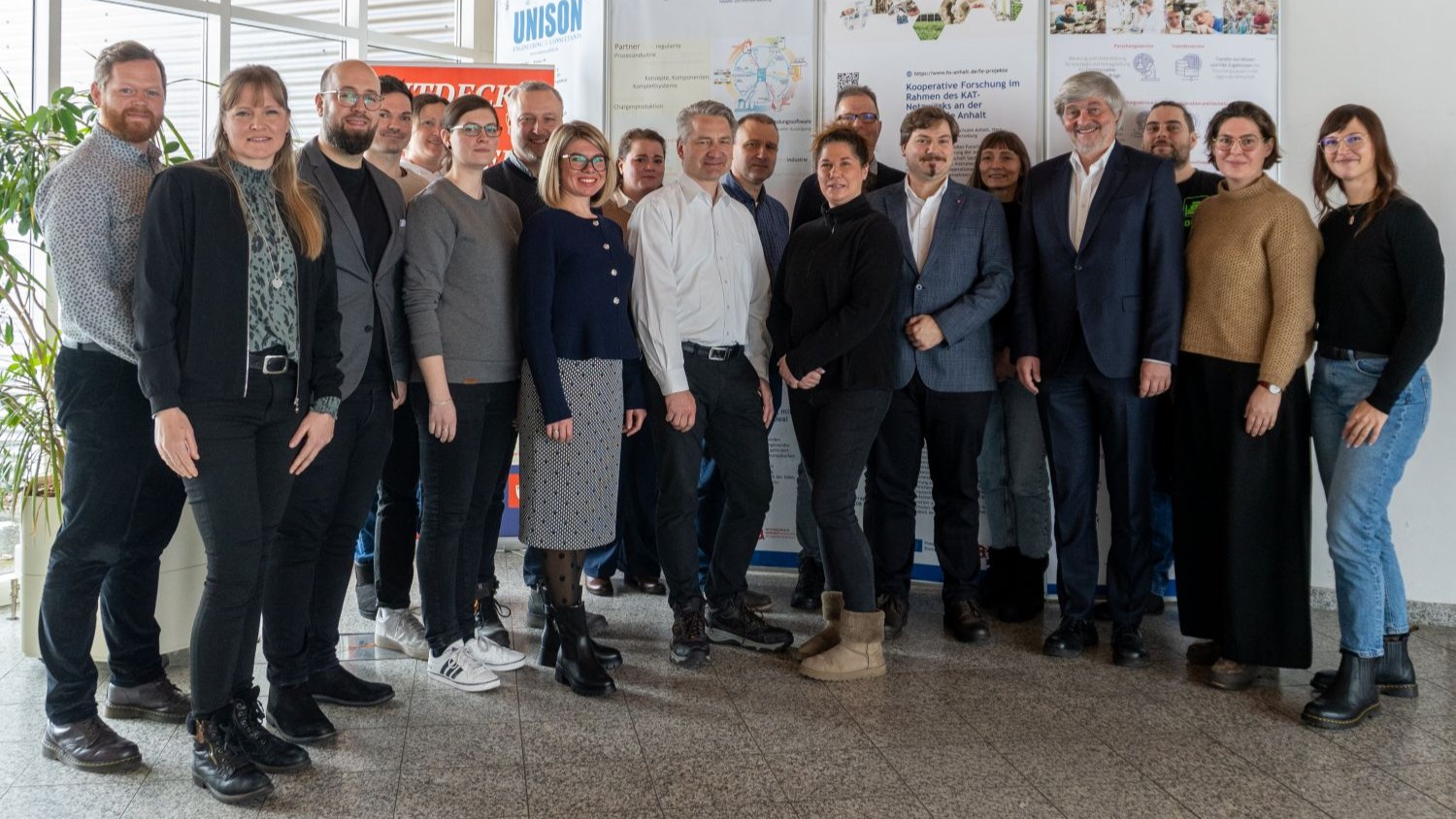 Gruppe von 18 Personen, stehend im Halbkreis, lächelnd für Gruppenfoto. Verschiedene Altersgruppen und Kleidungsstile, darunter Anzüge, Blusen und Jacken. Hintergrund zeigt eine Posterwand mit Informationsmaterial.