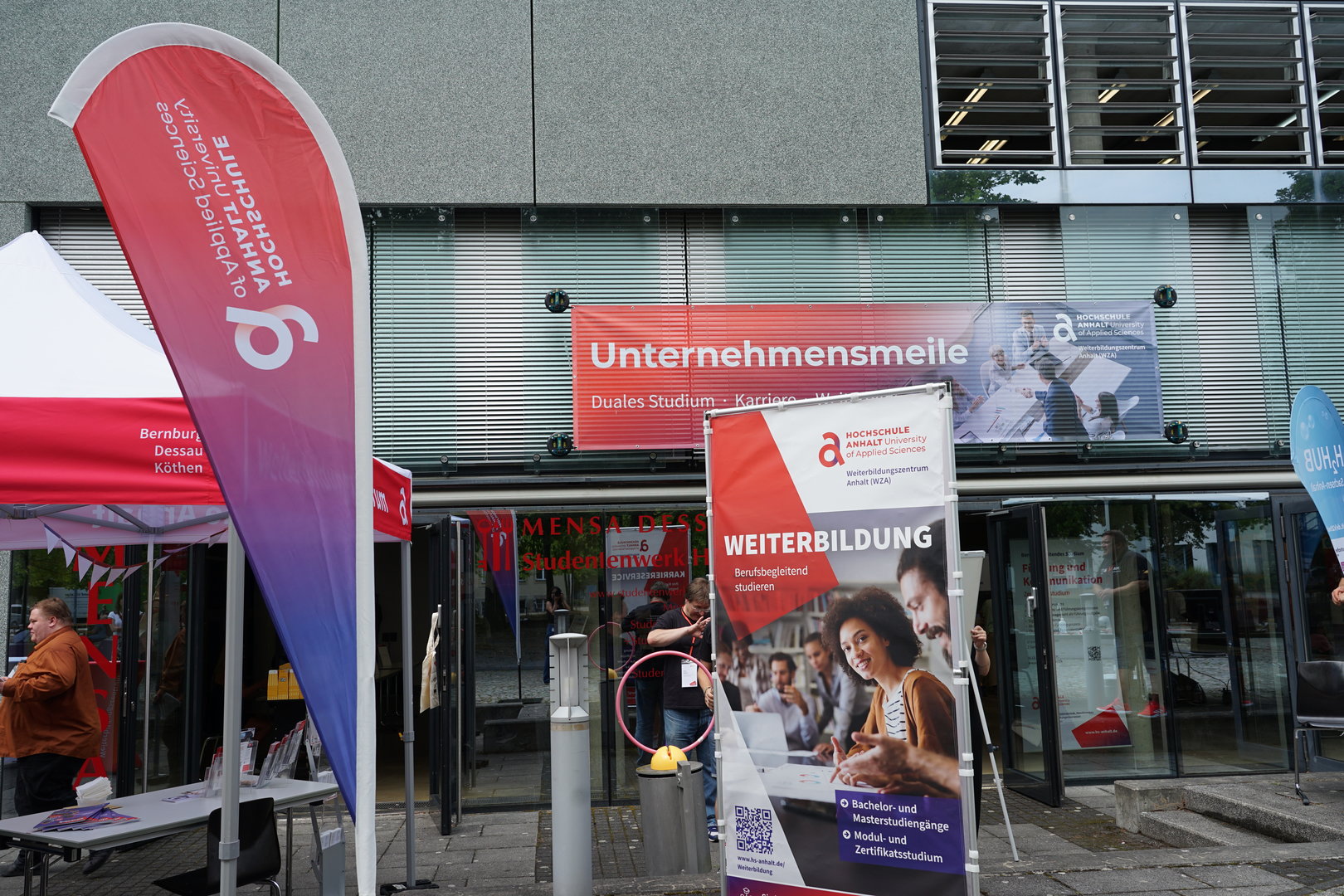 Ein Messeauftritt mit Infostand zur Unternehmensmeile, darunter ein Plakat für Weiterbildung mit dem Fokus auf „Duales Studium“ und „Ausbildung“. Eine Person ist auf dem Plakat beim Lernen mit Tablet zu sehen.