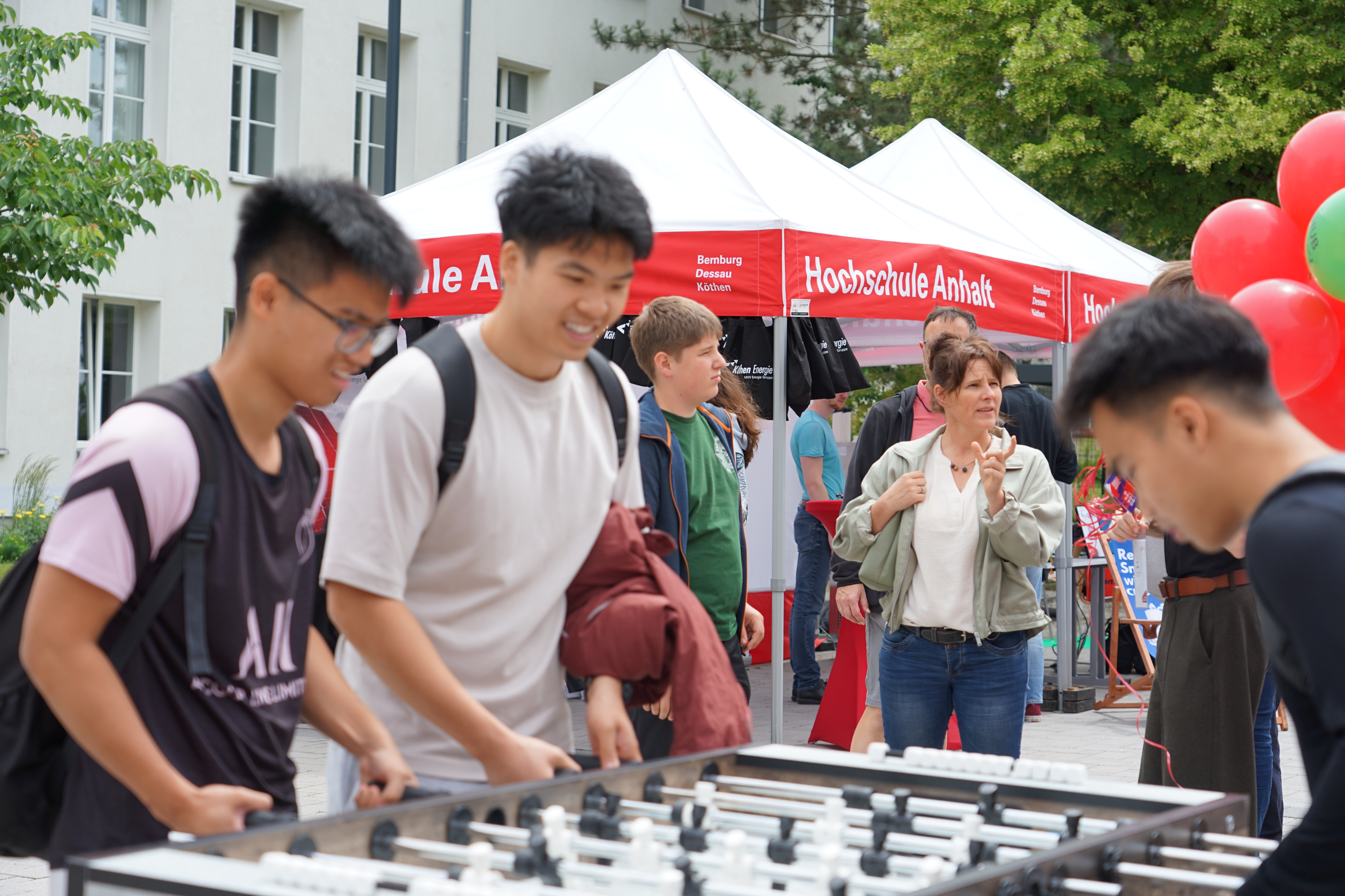 Zwei junge Männer spielen auf einem Foosballtisch, während im Hintergrund ein Infostand der Hochschule Anhalt mit mehreren Besuchern zu sehen ist.