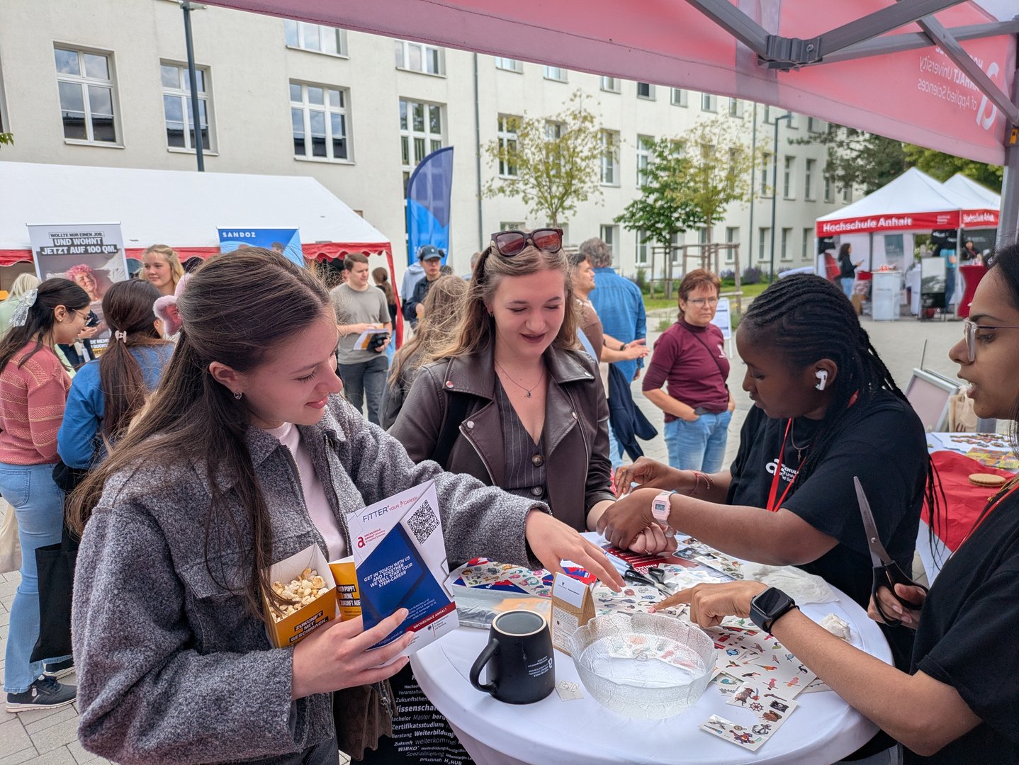 Drei Personen stehen an einem Infostand auf einer Veranstaltung. Eine Person mit grauer Jacke und eine Person mit brauner Jacke halten Flyer oder Broschüren, während eine dritte Person hinter einem Tisch mit Unterlagen und kleinen Gegenständen steht und etwas erklärt oder aushändigt.