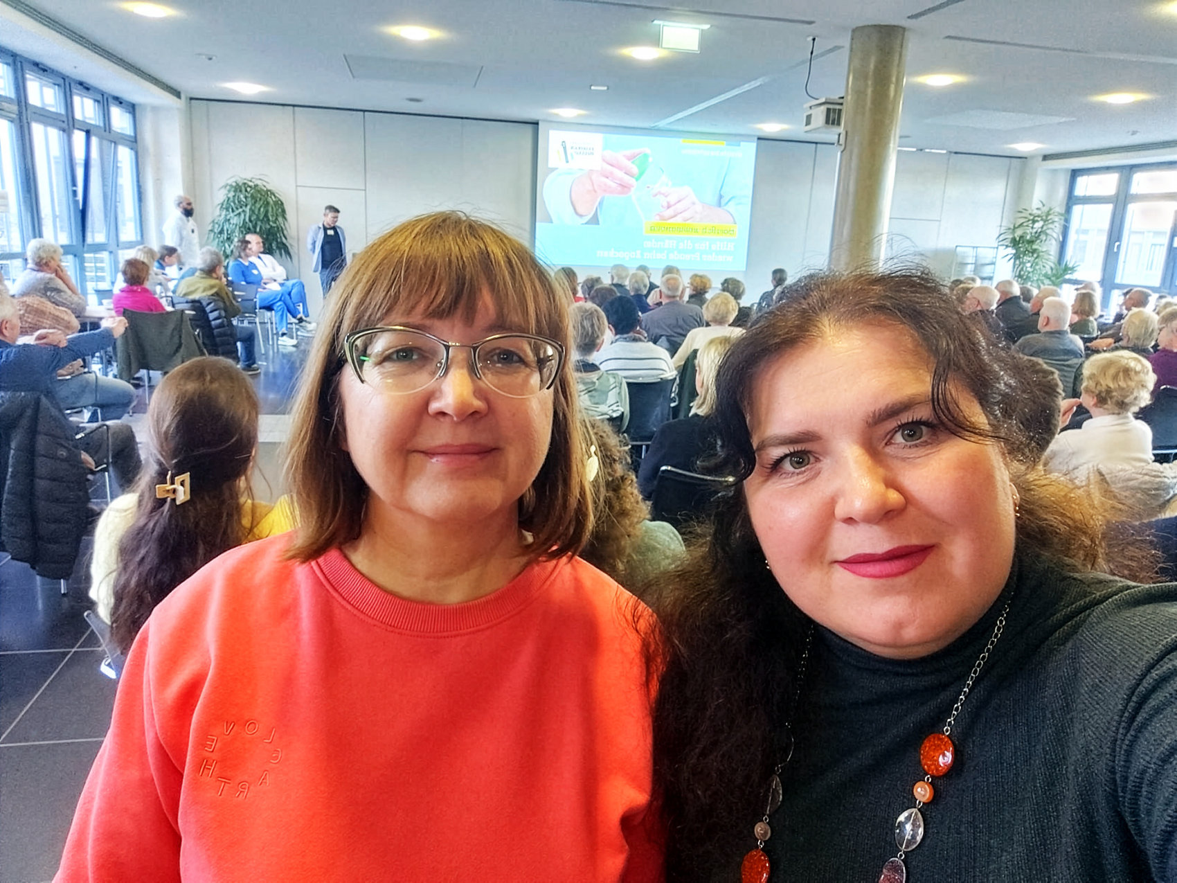 Zwei lächelnde Frauen sitzen vorne in einem gut besuchten Saal mit vielen Zuschauern, im Hintergrund eine große Leinwand mit einer Präsentation zu Herzthemen.