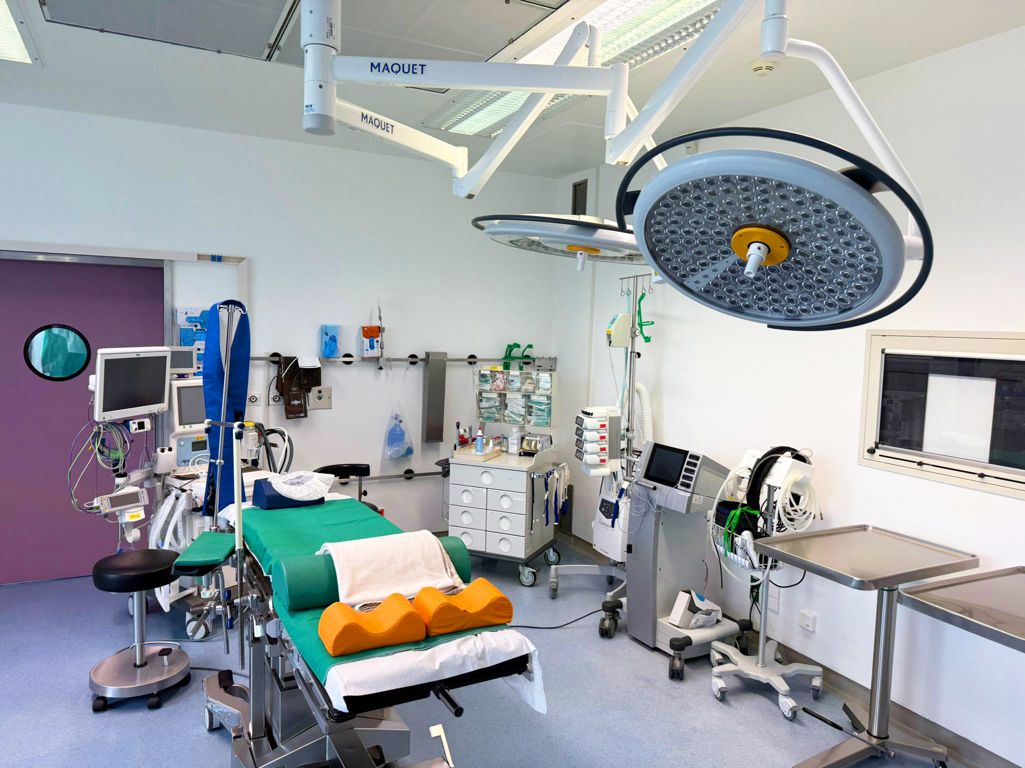 Moderner Operationssaal mit zentralem OP-Tisch, grünem OP-Tuch, zwei Operationsleuchten, medizinischen Geräten wie Monitoren und Infusionsständern sowie medizinischer Technik und Instrumenten an den Wänden.