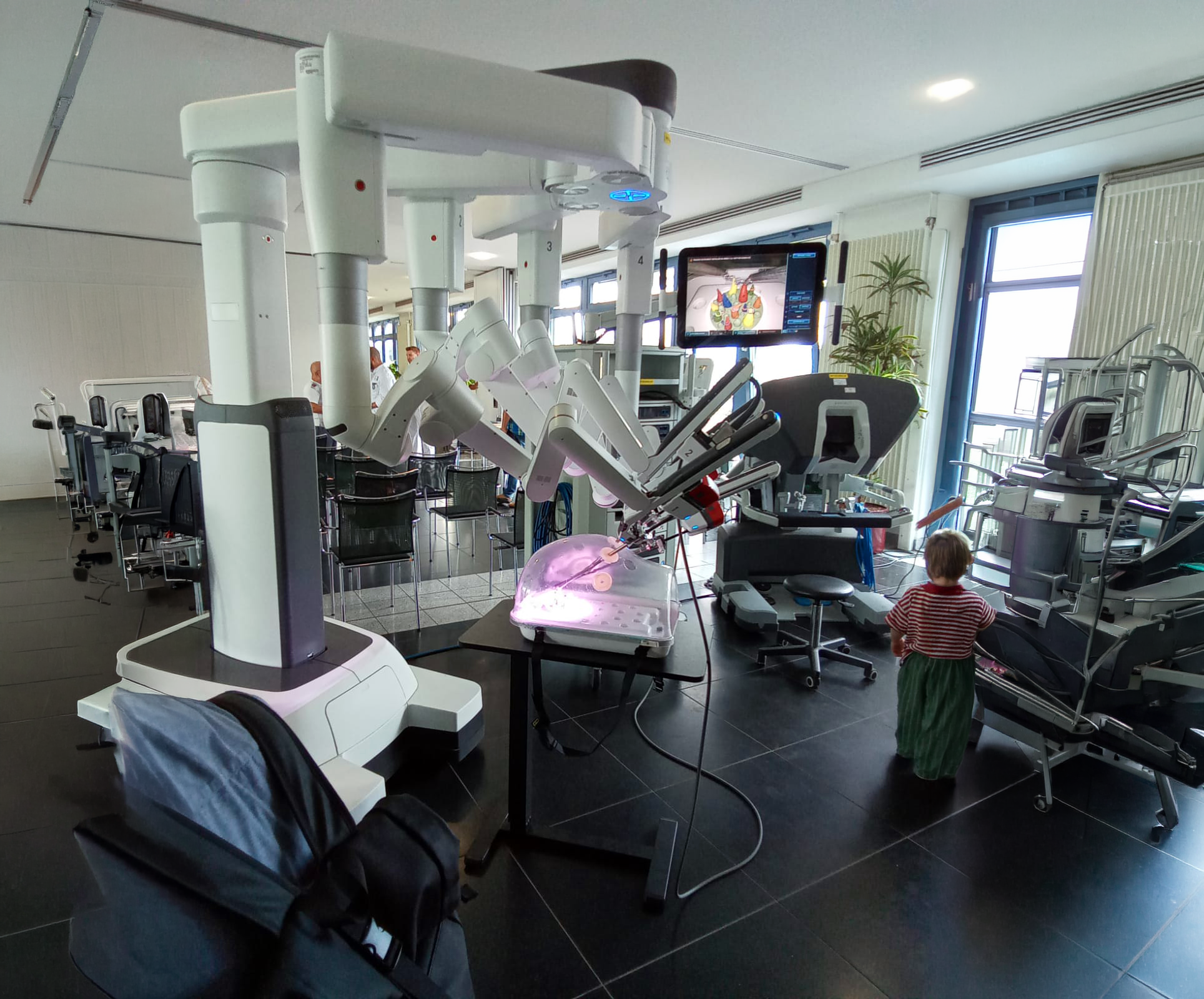 Moderner Operationssaal mit Roboterarm-System zur präzisen medizinischen Unterstützung, umgeben von medizinischen Geräten und einem Operationstisch. Ein Kind beobachtet die Szene im Hintergrund.