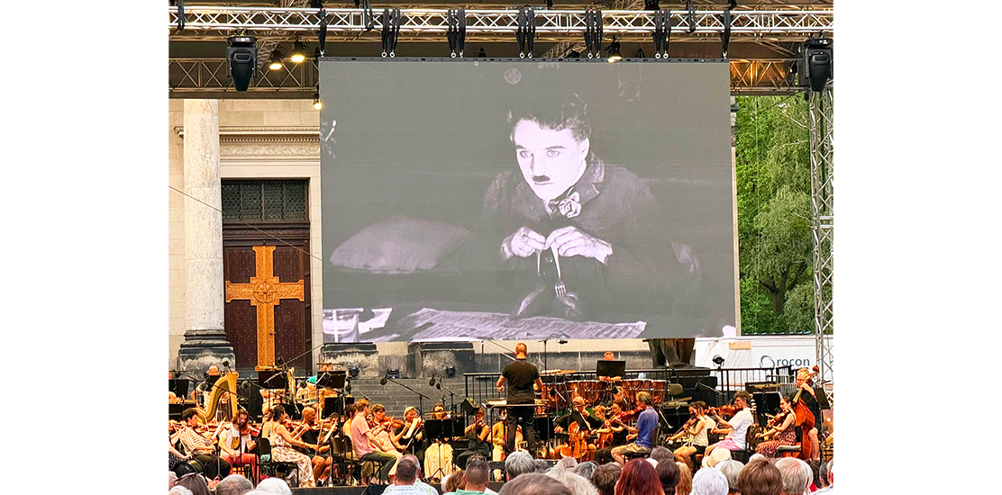 Großes Open-Air-Konzert mit Orchester und Chor vor einer Bühne, auf der ein Schwarz-Weiß-Film von Charlie Chaplin mit ihm in einer traurigen Pose beim Geigenspiel projiziert wird.