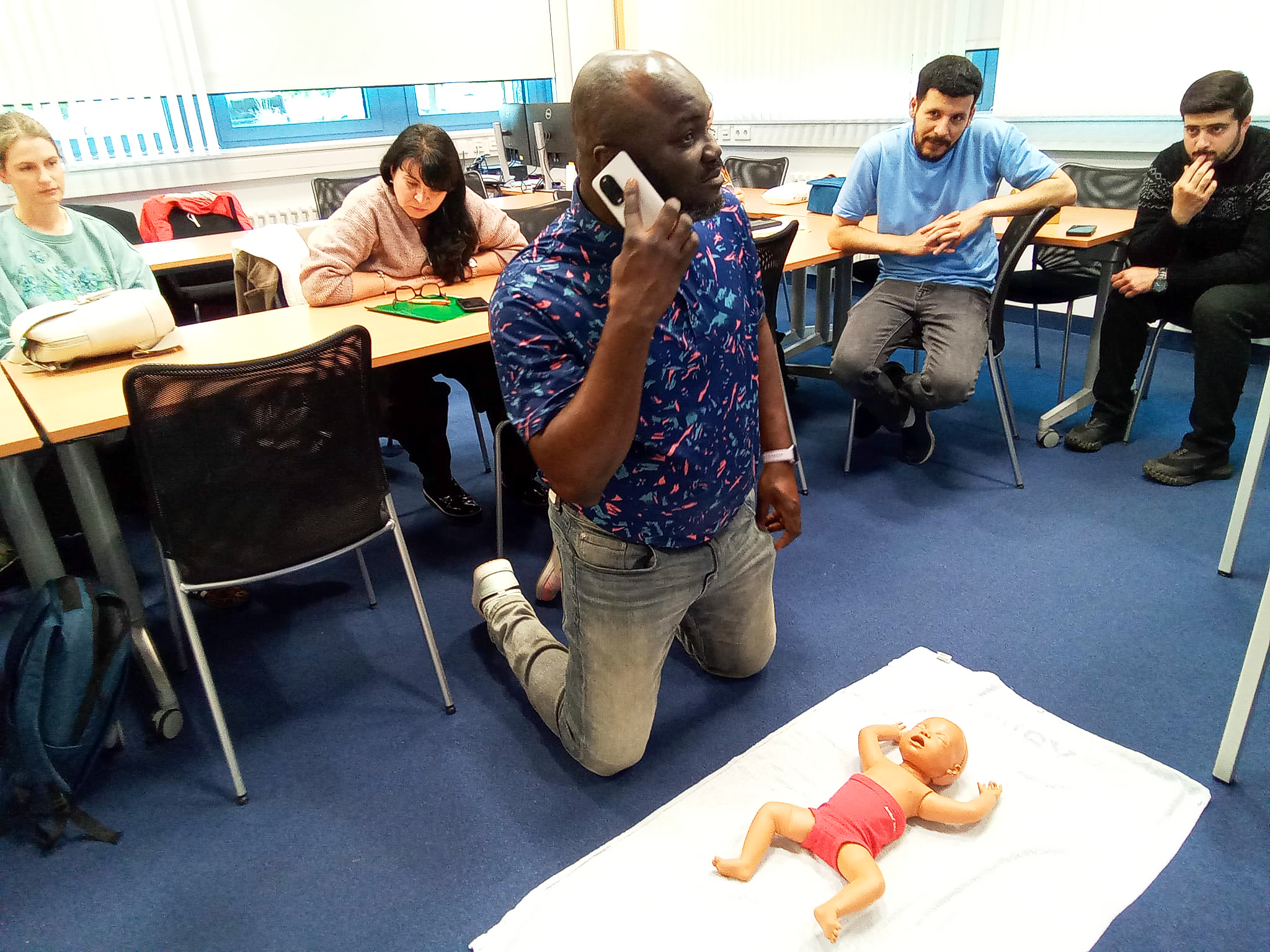 Ein Mann kniet vor einer Gruppe von Menschen in einem Raum und telefoniert, während er eine Puppe in Babykleidung auf einem Tuch hält. Die Gruppe beobachtet die Situation.