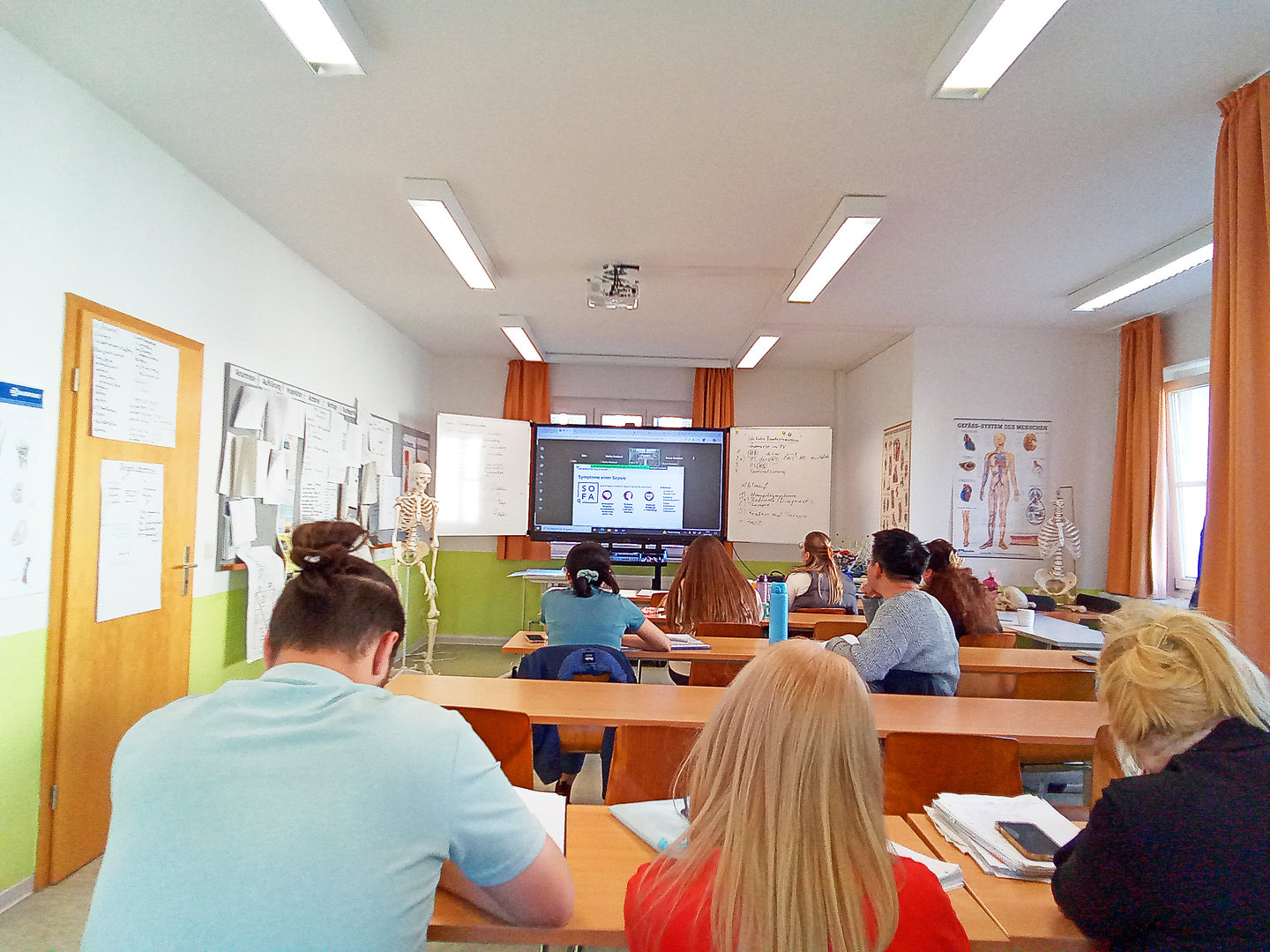 Ein Klassenzimmer mit mehreren Schülern, die einem Unterricht folgen. Vorne steht eine Lehrkraft, während am Whiteboard und auf einem Bildschirm medizinische Anatomie, insbesondere ein Skelett, dargestellt wird.