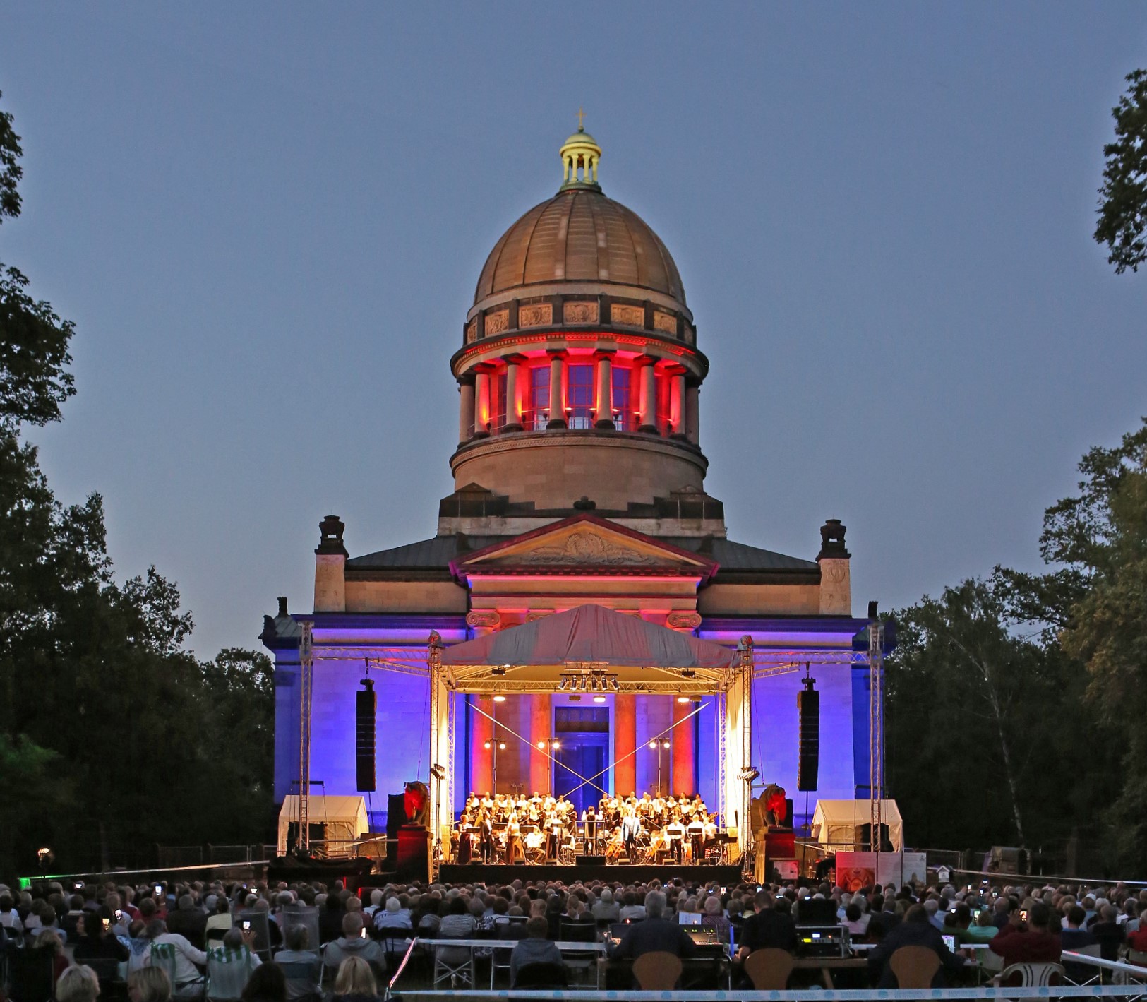 Abendkonzert vor einem klassizistischen Kuppelbau mit Säulenportikus, beleuchtet in rot und violett. Ein großes Orchester spielt auf der Bühne, während ein Publikum auf Stühlen im Freien zuhört.