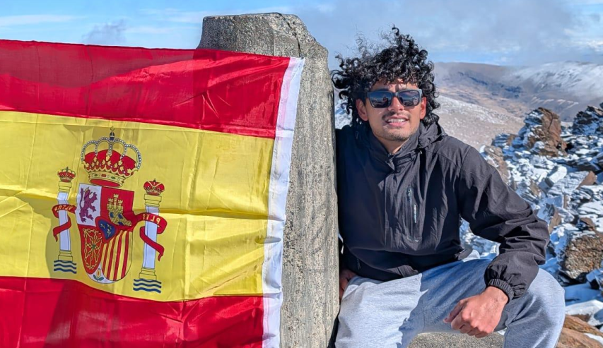 Ein Mann sitzt auf einem Felsen in bergiger, schneebedeckter Landschaft und hält die spanische Flagge mit dem Wappen in der Mitte.