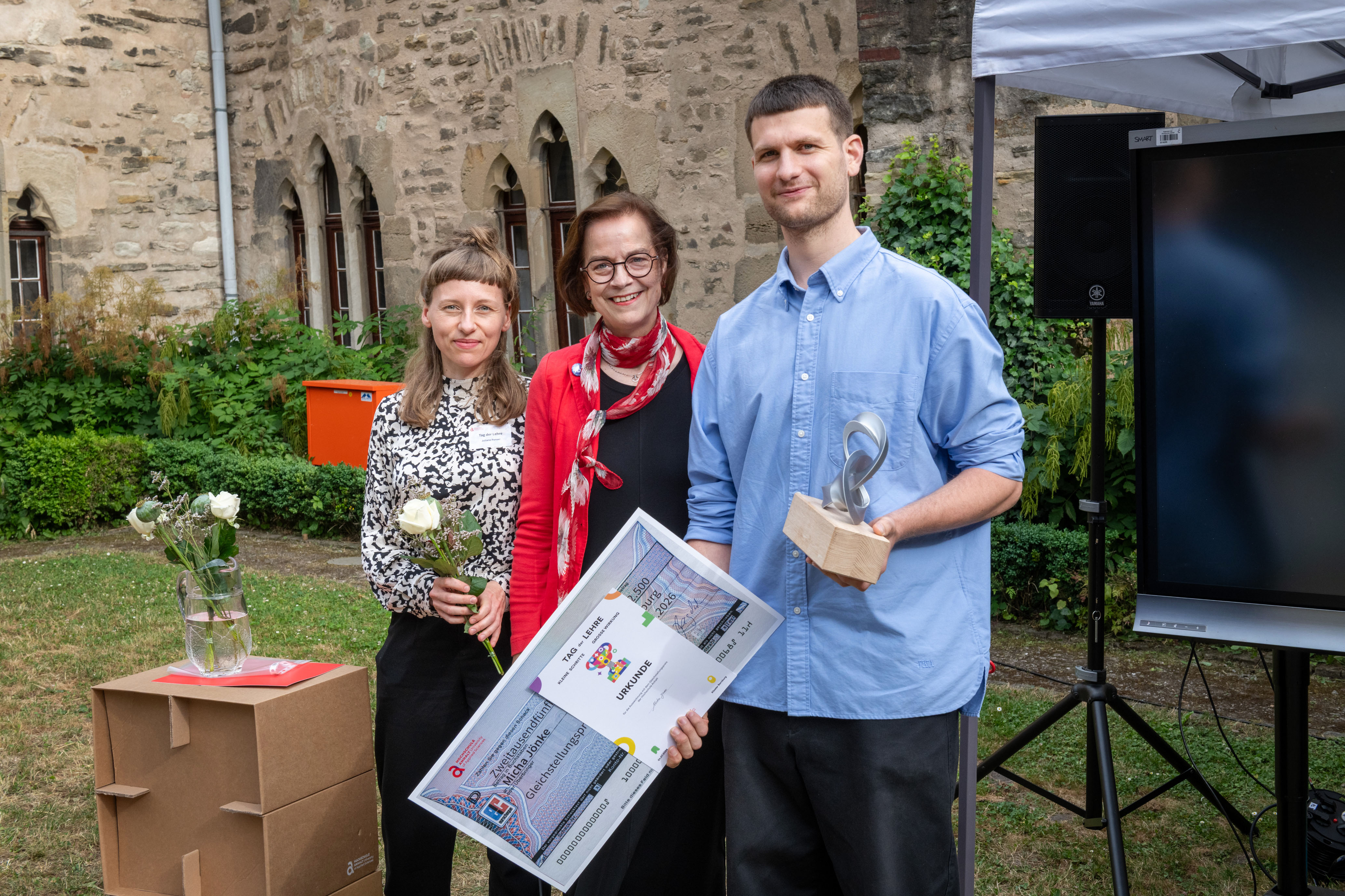 Drei Personen vor historischer Mauer: Eine Frau hält eine Urkunde mit Logo „TAO – LEMRE“ und Blumen, eine zweite Frau steht lächelnd daneben, ein Mann hält eine Trophäe in Form eines stilisierten Buches. Szene wirkt wie eine Preisverleihung.