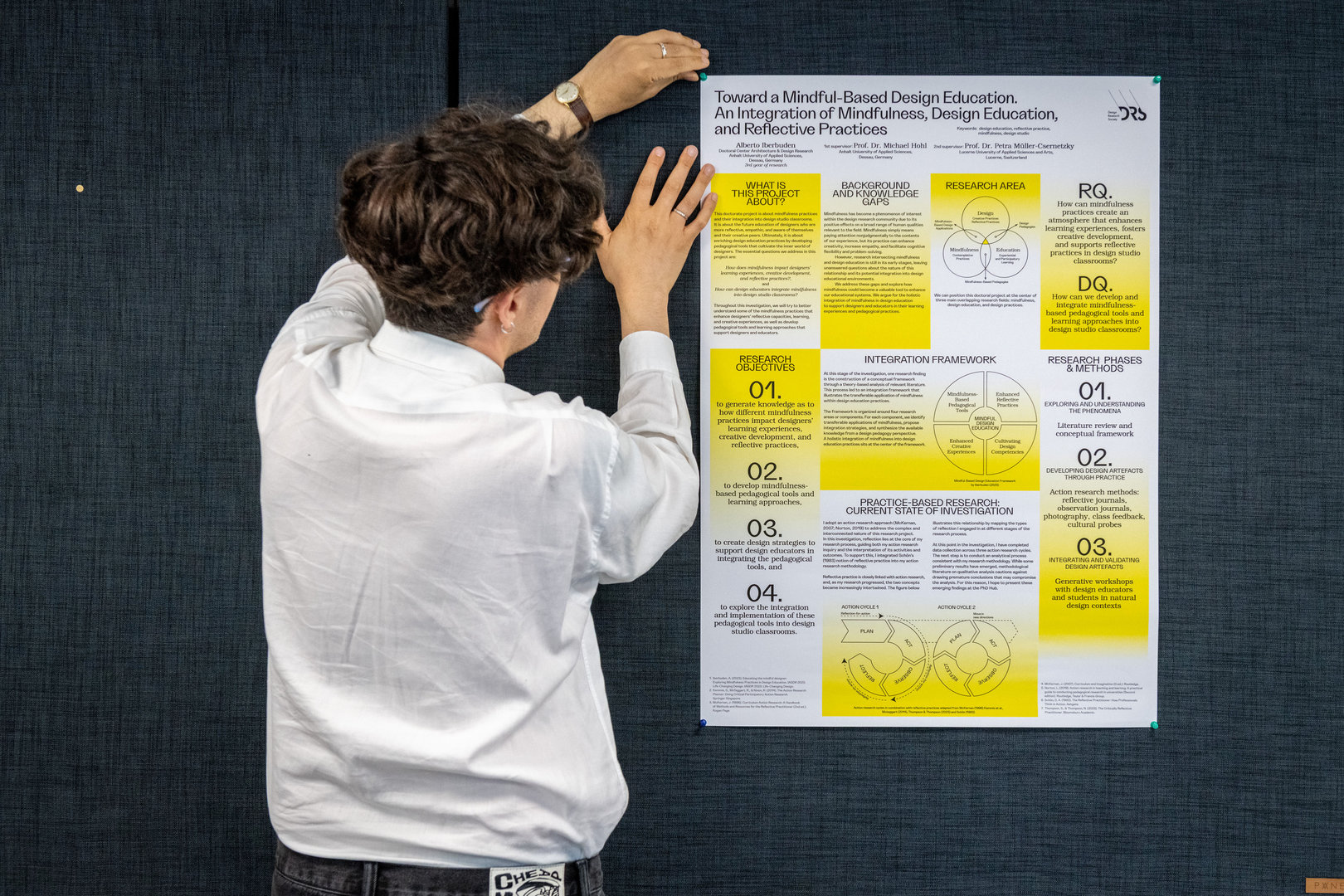 Eine Person berührt mit beiden Händen eine Präsentation zum Thema „Toward a Mindful-Based Design Education“, die sich mit der Verbindung von Achtsamkeit, Designausbildung und reflektierenden Praktiken beschäftigt. Dargestellt sind Forschungsfragen, Ziele und Methoden zur Integration von Achtsamkeit in Designprozesse.