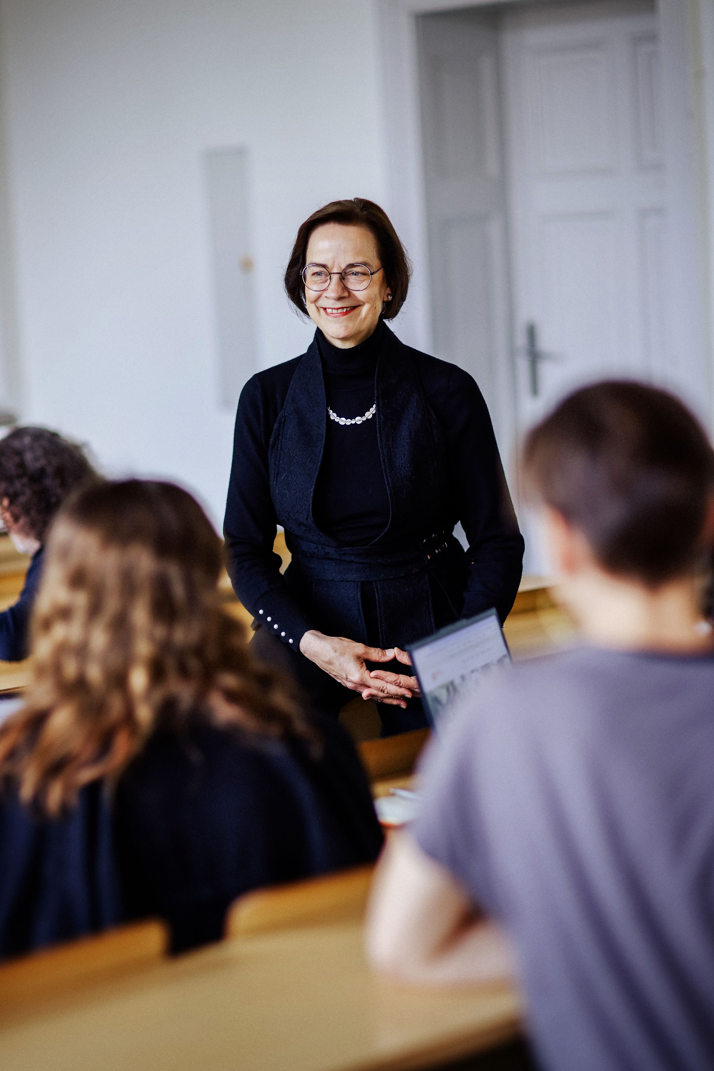 Eine lächelnde Dozentin steht vor einer Gruppe von Studierenden im Hörsaal und hält ein Tablet, während sie eine Präsentation abhält.