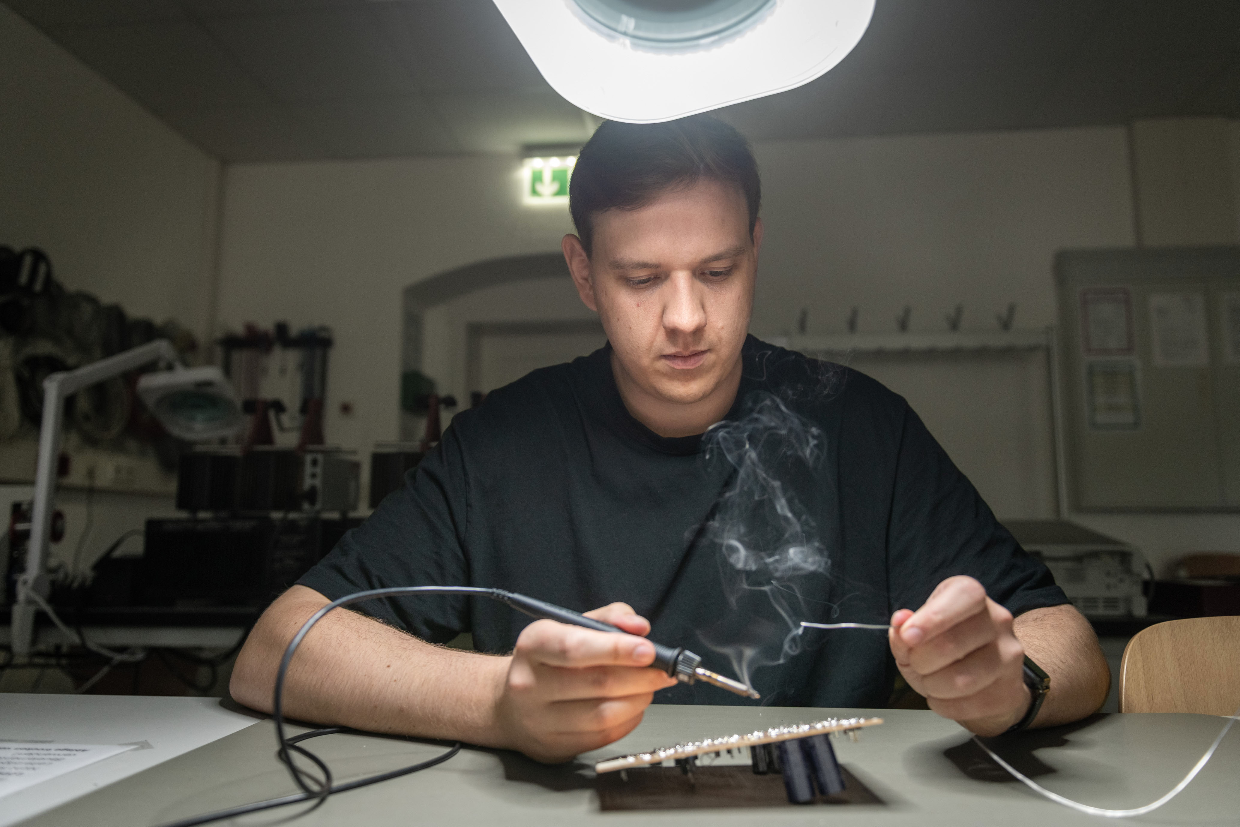 Eine Person lötet konzentriert an einer elektronischen Platine, während leichter Rauch aus dem Lötkolben aufsteigt. Im Hintergrund ist ein Arbeitsplatz mit Werkzeugen und Laborausstattung zu erkennen.