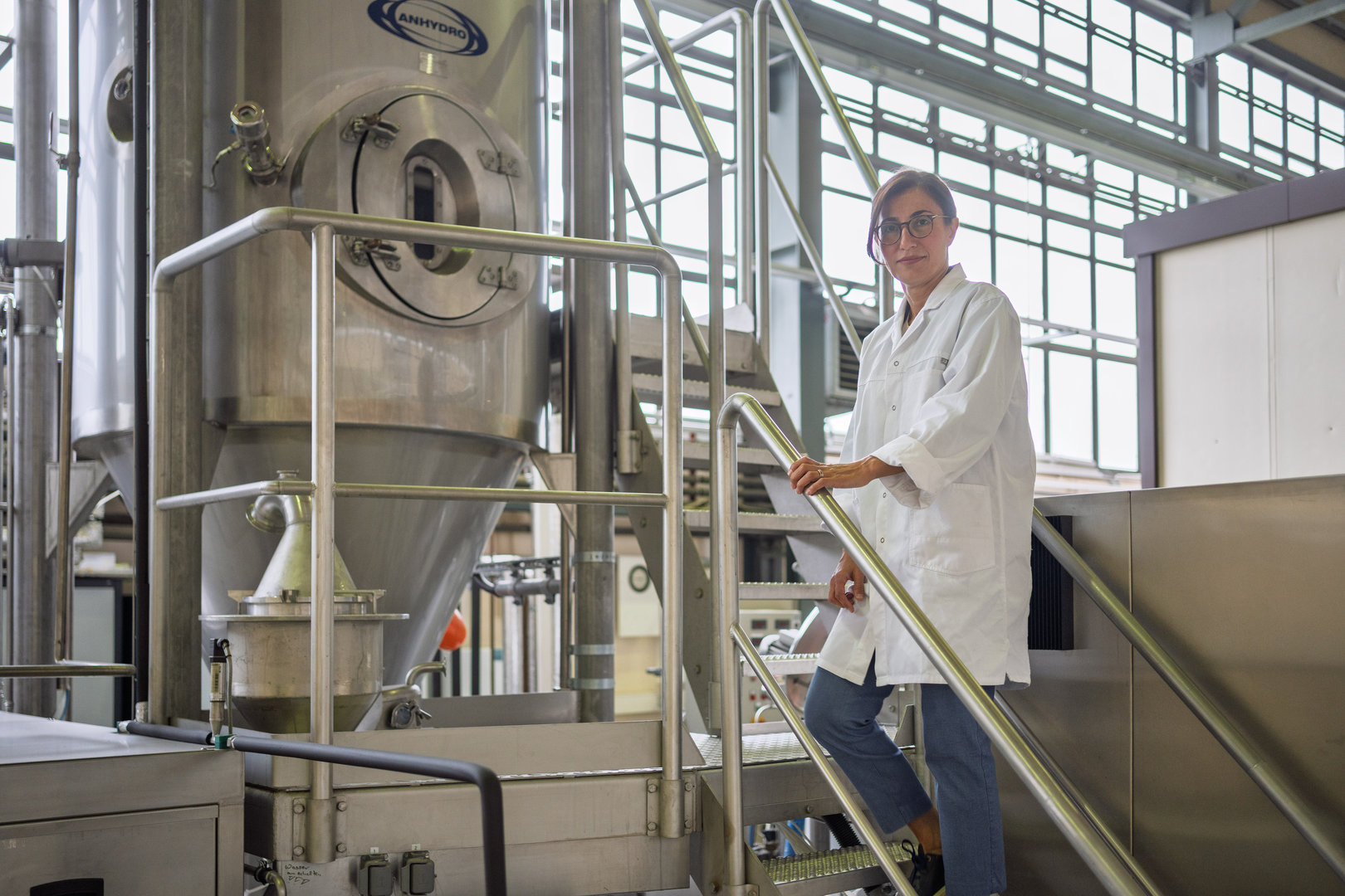 Eine Person in weißem Laborkittel steht lächelnd auf einer Metalltreppe in einer modernen Produktionsanlage mit großen Edelstahltanks und Rohrleitungen, vermutlich in einer Brauerei oder Lebensmittelproduktion.