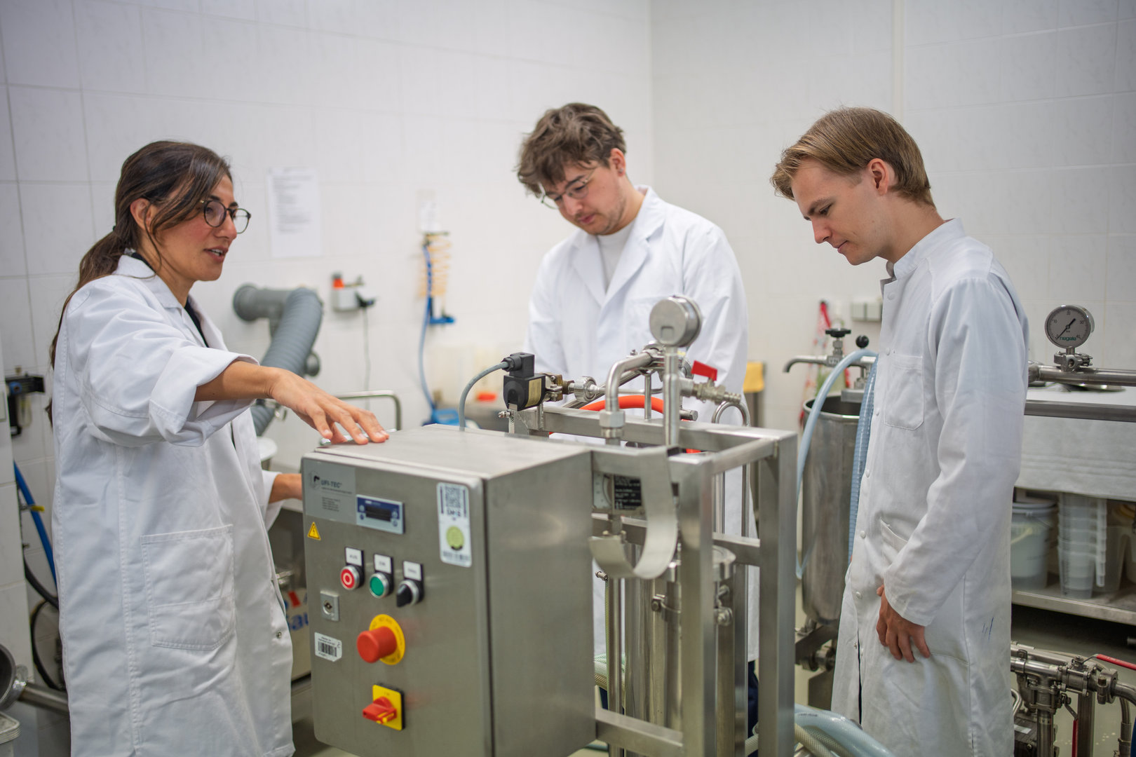 Drei Personen in Laborkitteln stehen vor einer industriellen Maschine mit Bedienelementen und Anzeigen, die anscheinend eine Flüssigkeit verarbeitet. Sie scheinen eine Bedienungsanleitung oder ein Verfahren zu besprechen.