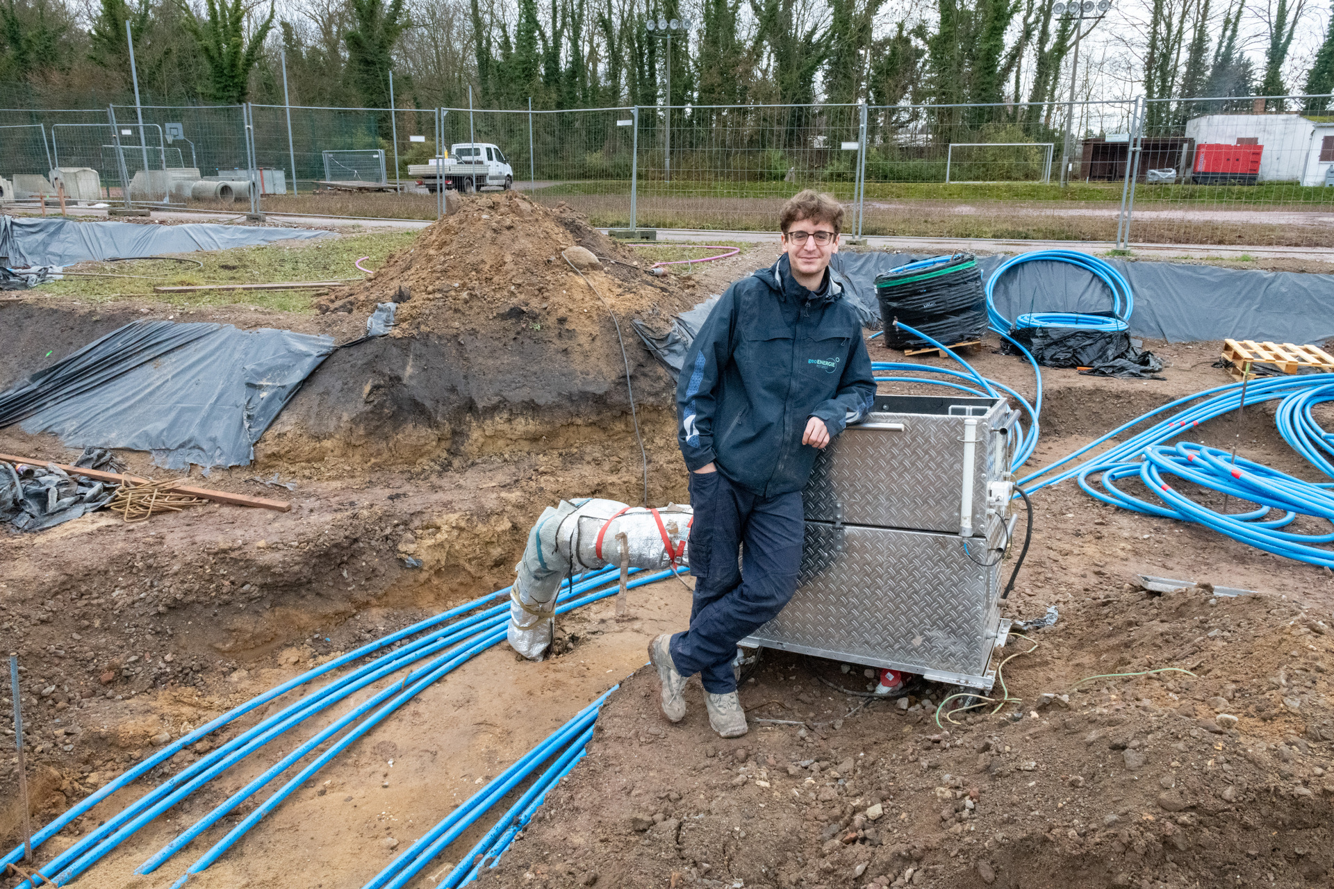 Ein Mann steht auf einer Baustelle neben einer Maschine mit blauen Schläuchen. Er trägt eine dunkle Jacke und Jeans. Im Hintergrund sind Erde, Planen und ein Zaun zu sehen. Die Umgebung wirkt wie eine Baustelle oder ein Industriegebiet.