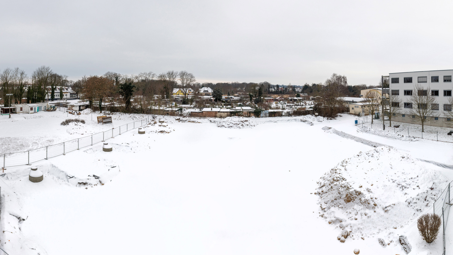 Ein verschneites, leeres Grundstück mit einem Zaun und Betonpfosten. Im Hintergrund sind Wohnhäuser und Bäume zu sehen. Die Umgebung wirkt ruhig und winterlich. Der Himmel ist bewölkt.