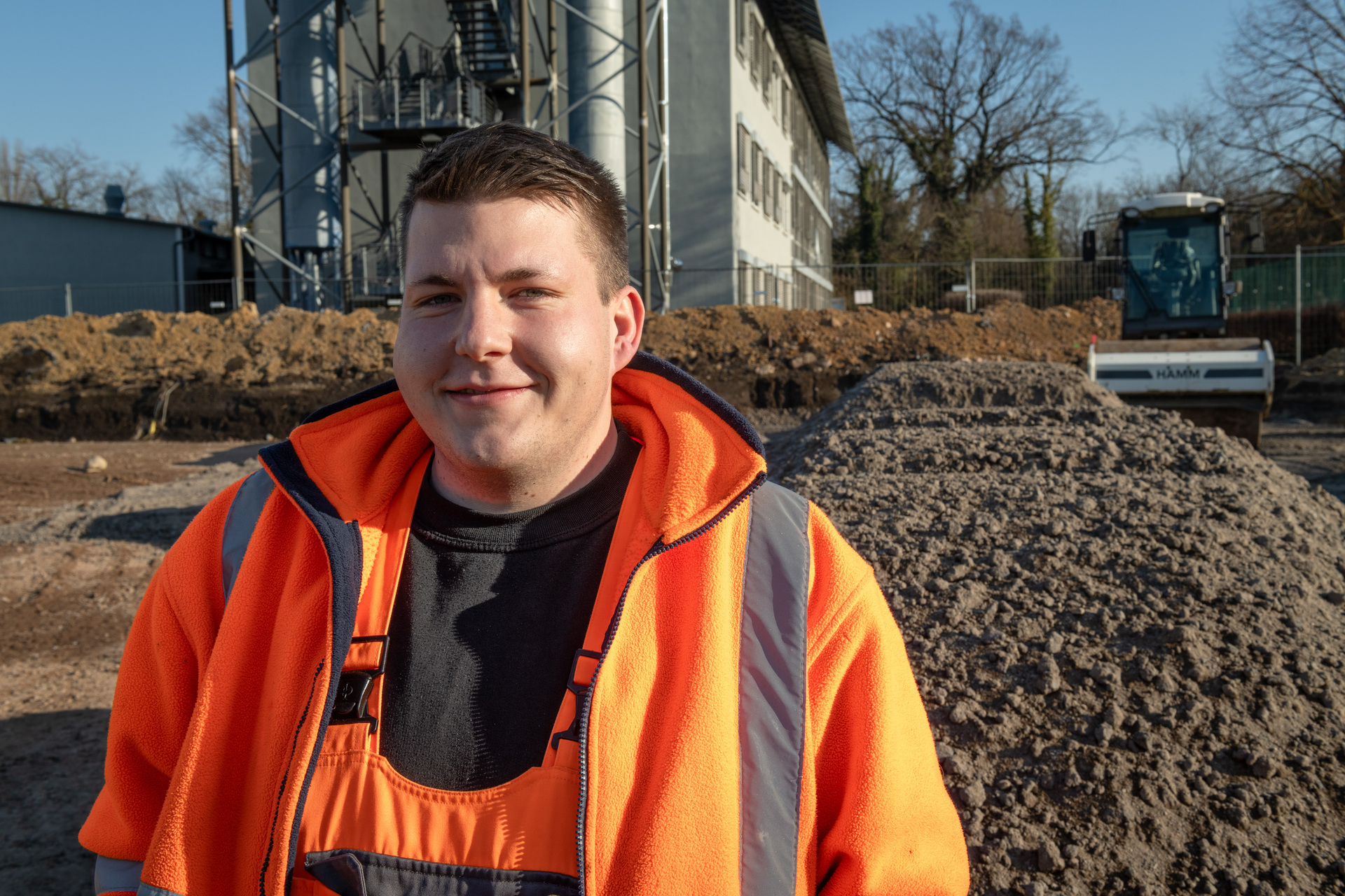 Ein Arbeiter in einer Konstruktionsumgebung lächelt freundlich. Er trägt eine hellorange Warnjacke über einem dunklen Shirt und steht vor einem großen Materialhaufen, vermutlich Sand oder Erde. Die Umgebung wirkt nach industriellem oder Baustellengelände. Er scheint sich sicher und zufrieden auf seiner Arbeitsstelle zu befinden.
