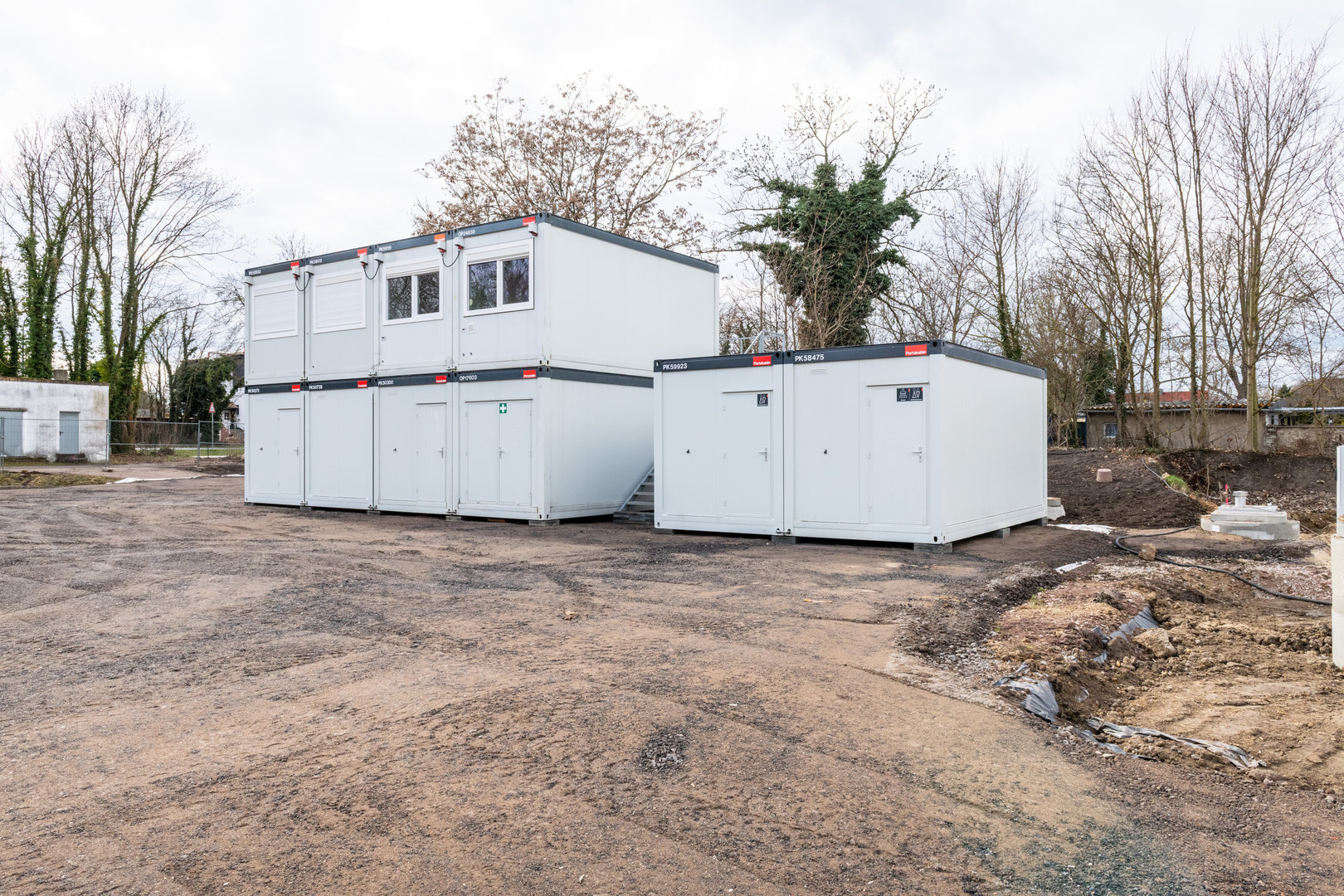 Zwei weiße Container, die aneinandergebaut sind, stehen auf einem unbefestigten Platz. Die Container haben Türen mit Schildern und Fenster an der Vorderseite, vermutlich als Technik- oder Transformatorenstation genutzt.